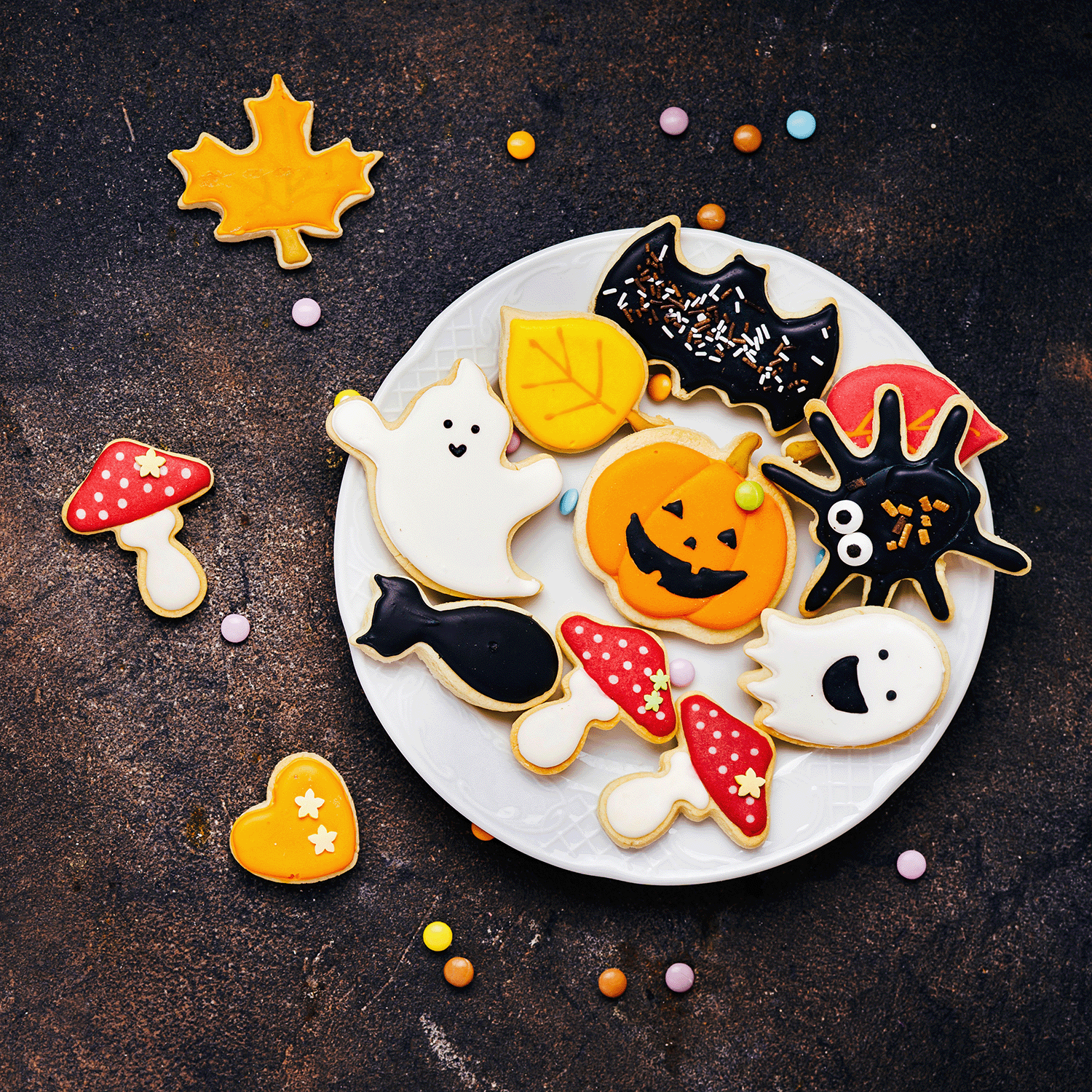 A plate of Halloween-themed cookies shaped like ghosts, a pumpkin, a spider, and a bat is surrounded by leaf cookies, mushrooms, hearts, and scattered colorful candies. Enhance the spooky ambiance with a Monster Cookies Long-Lasting Scented Jar Candle (12 oz) from Tuscany Candle® SEASONAL for an extra touch of eerie delight.