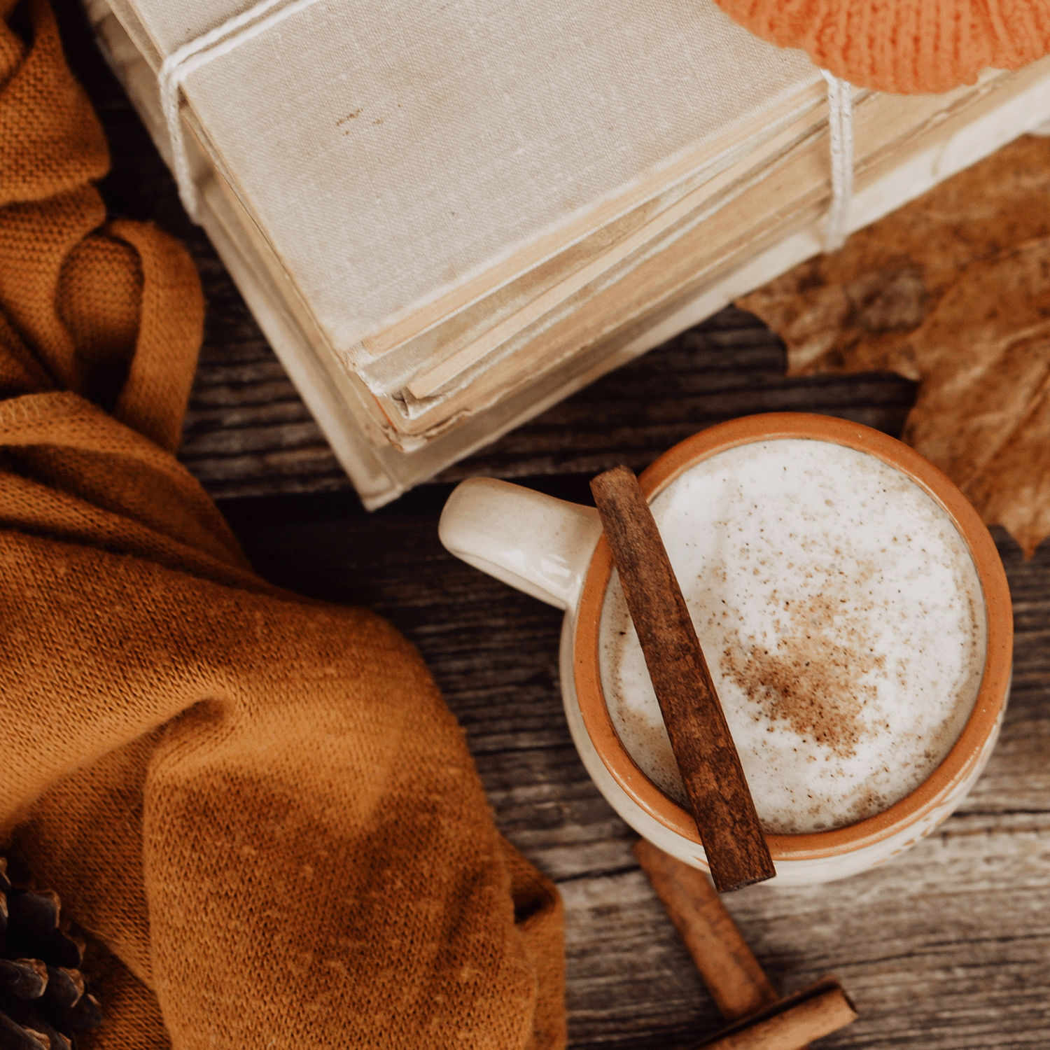 A jar of Tuscany Candle® SEASONAL's Creamy Pumpkin Latte Long-Lasting Scented Jar Candle (12 oz), a stack of books, an orange sweater, a pinecone, and a dried leaf on a wooden table.