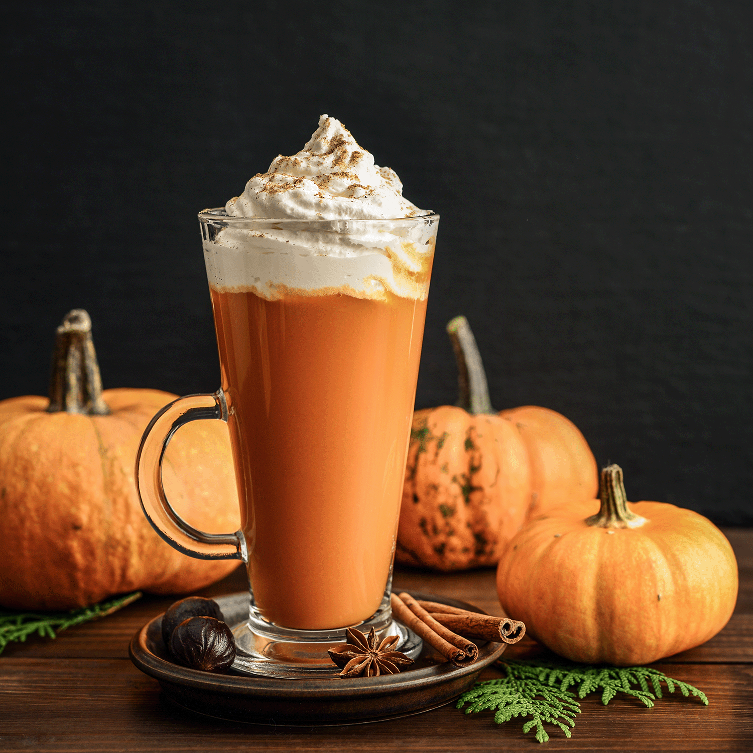 A glass mug of spiced pumpkin latte, topped with whipped cream and sprinkled with spices, sits on a wooden table alongside small pumpkins, cinnamon sticks, and star anise. Nearby, a 12 oz Tuscany Candle® SEASONAL Creamy Pumpkin Latte Long-Lasting Scented Jar Candle enhances the cozy autumn ambiance.