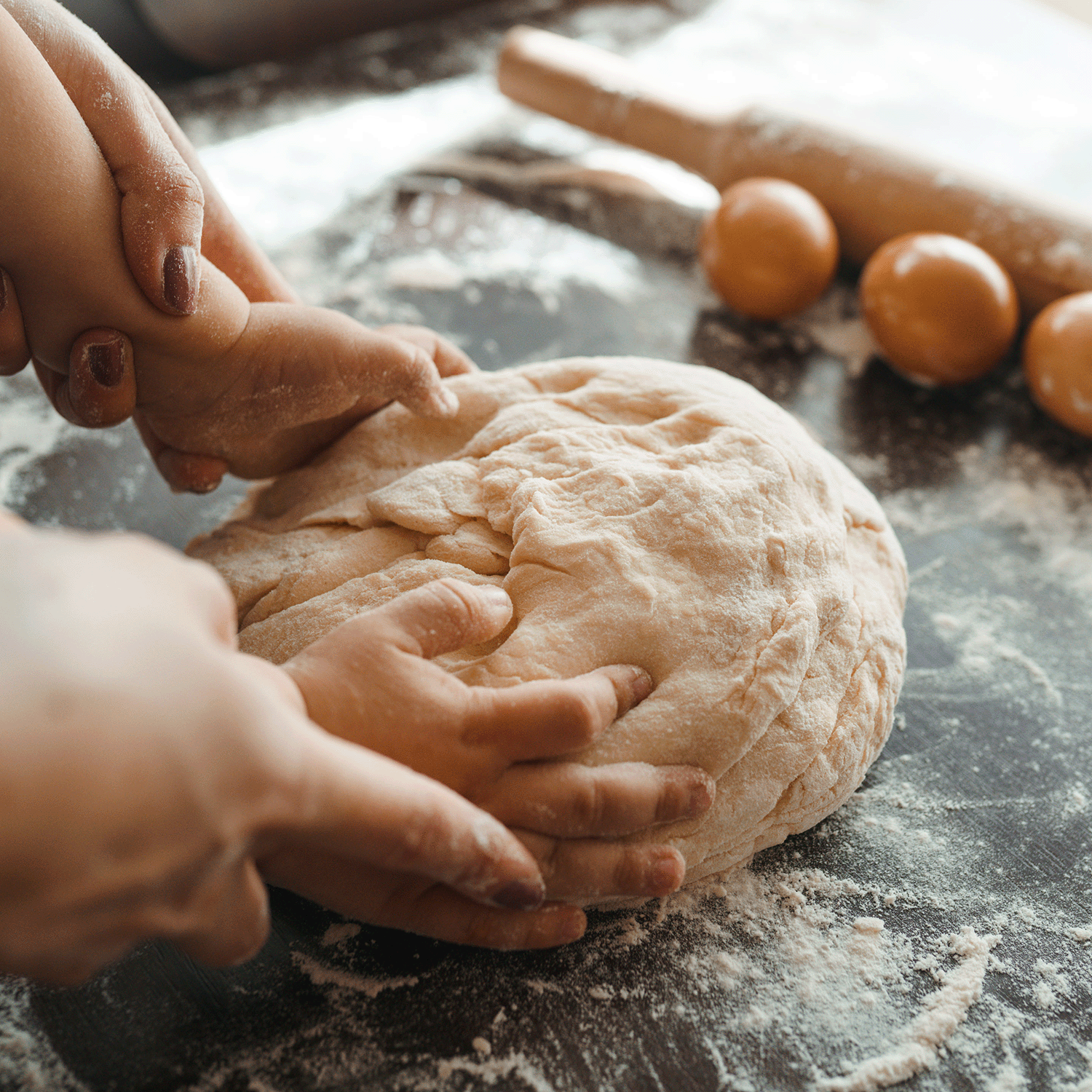 Hands kneading dough on a floured surface, with a rolling pin and eggs visible in the background. The warm, nutty fragrance from a nearby Pistachio Almond Long-Lasting Scented Jar Candle (18 oz) by Tuscany Candle® SEASONAL, part of the Fall Collection, creates an inviting atmosphere perfect for baking.