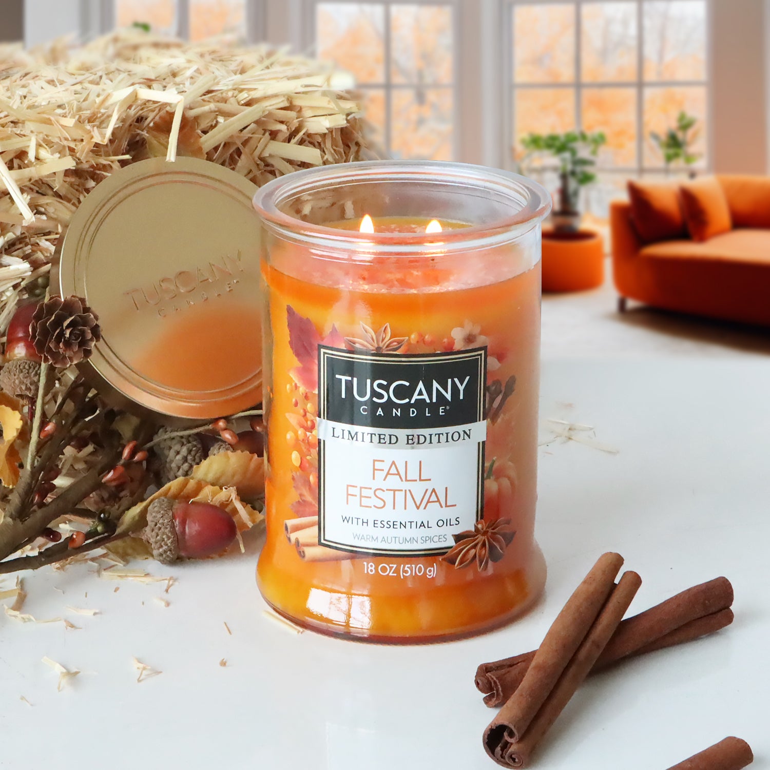 A Tuscany Candle® SEASONAL "Fall Festival Long-Lasting Scented Jar Candle (18 oz)" burns on a white surface, accompanied by cinnamon sticks, straw, and autumn decor in the background. This candle is part of a limited-run collection and fills the room with warm autumn spices. An orange sofa and windows showcasing a fall view are visible in the scene.