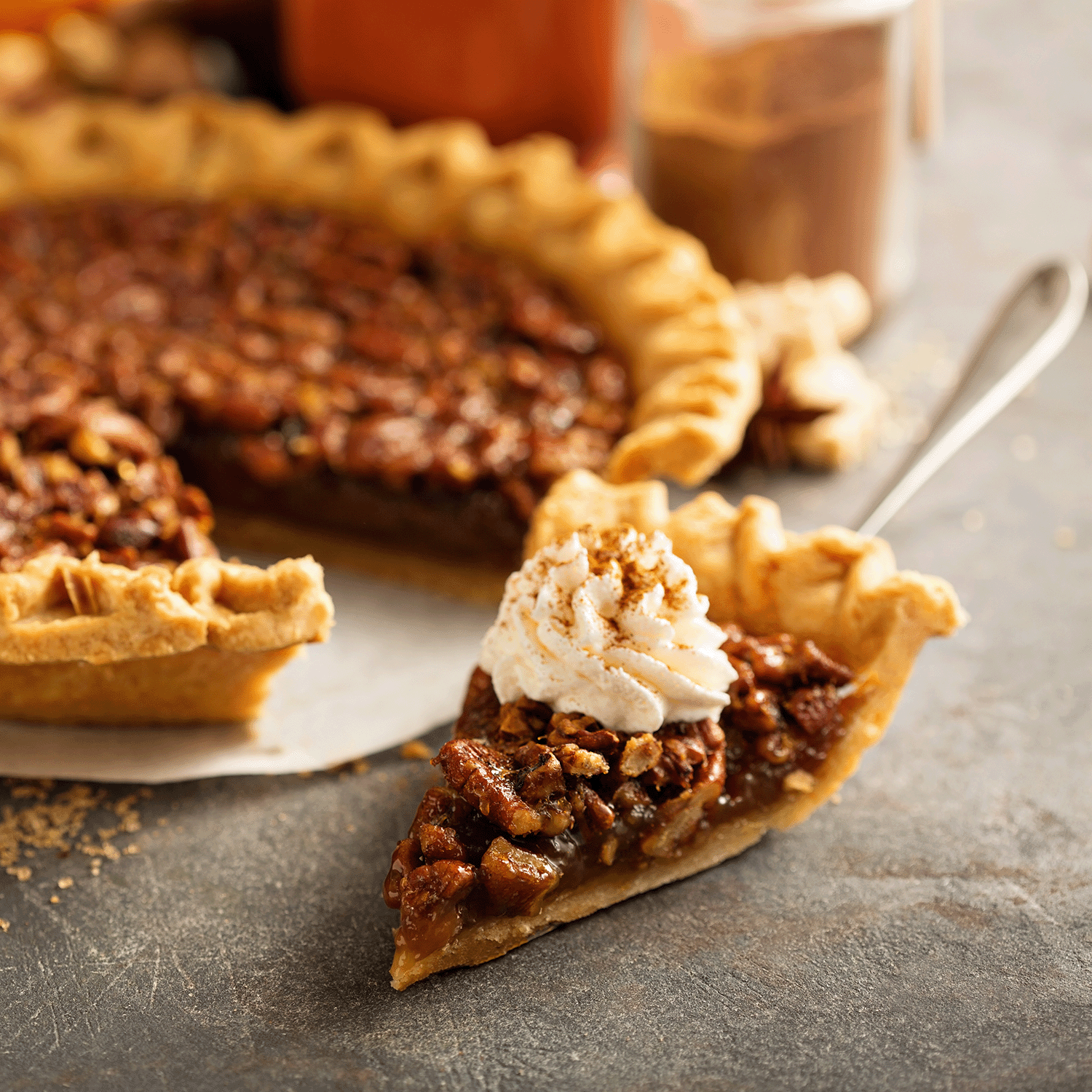 A slice of Pumpkin Pecan pie, garnished with whipped cream and a drizzle of maple-infused caramel, is served against the backdrop of an 18 oz Tuscany Candle® SEASONAL Pumpkin Pecan Long-Lasting Scented Jar Candle on a gray surface.