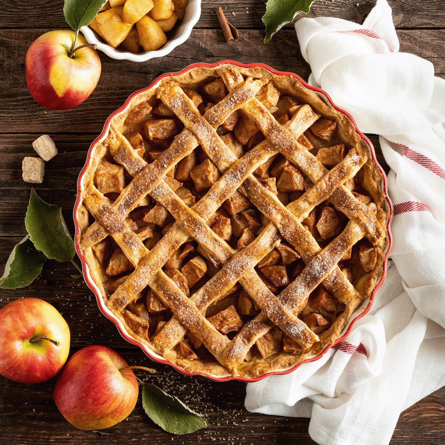 A freshly baked lattice-topped apple pie in a round dish on a wooden surface exudes an autumn fragrance, accompanied by whole apples, a small bowl of diced brown sugared apples, and a white cloth with red stripes. Nearby, the Give Thanks Long-Lasting Scented Jar Candle (18 oz) from the Tuscany Candle® SEASONAL Gather Together Collection adds warmth to the cozy scene.