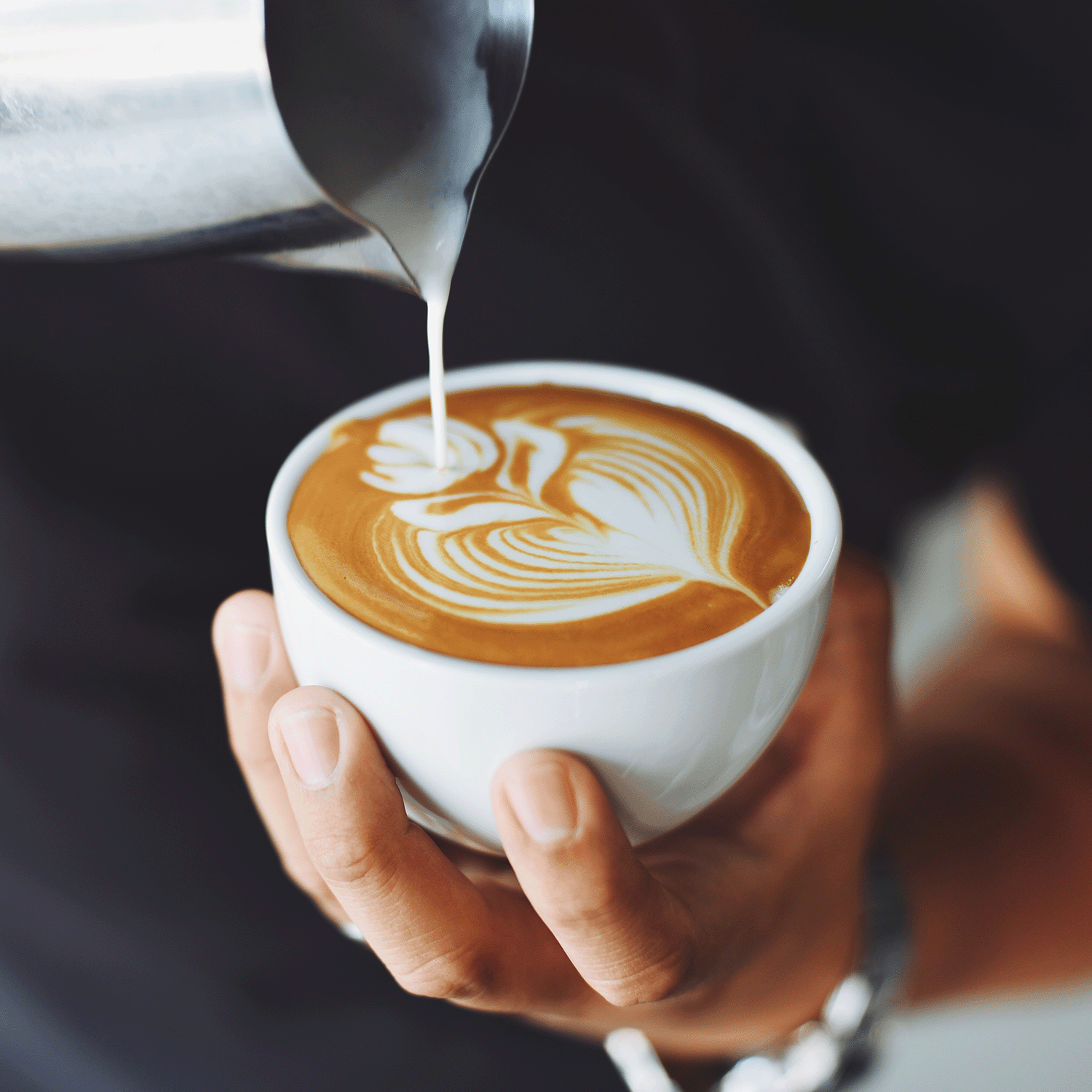 Close-up of a person pouring steamed milk into a coffee cup, creating latte art in the shape of a leaf, perfectly complementing the cozy vibes of the Autumn Flannels Collection's Pumpkin Spice Everything Long-Lasting Scented Jar Candle (12 oz) by Tuscany Candle® SEASONAL.