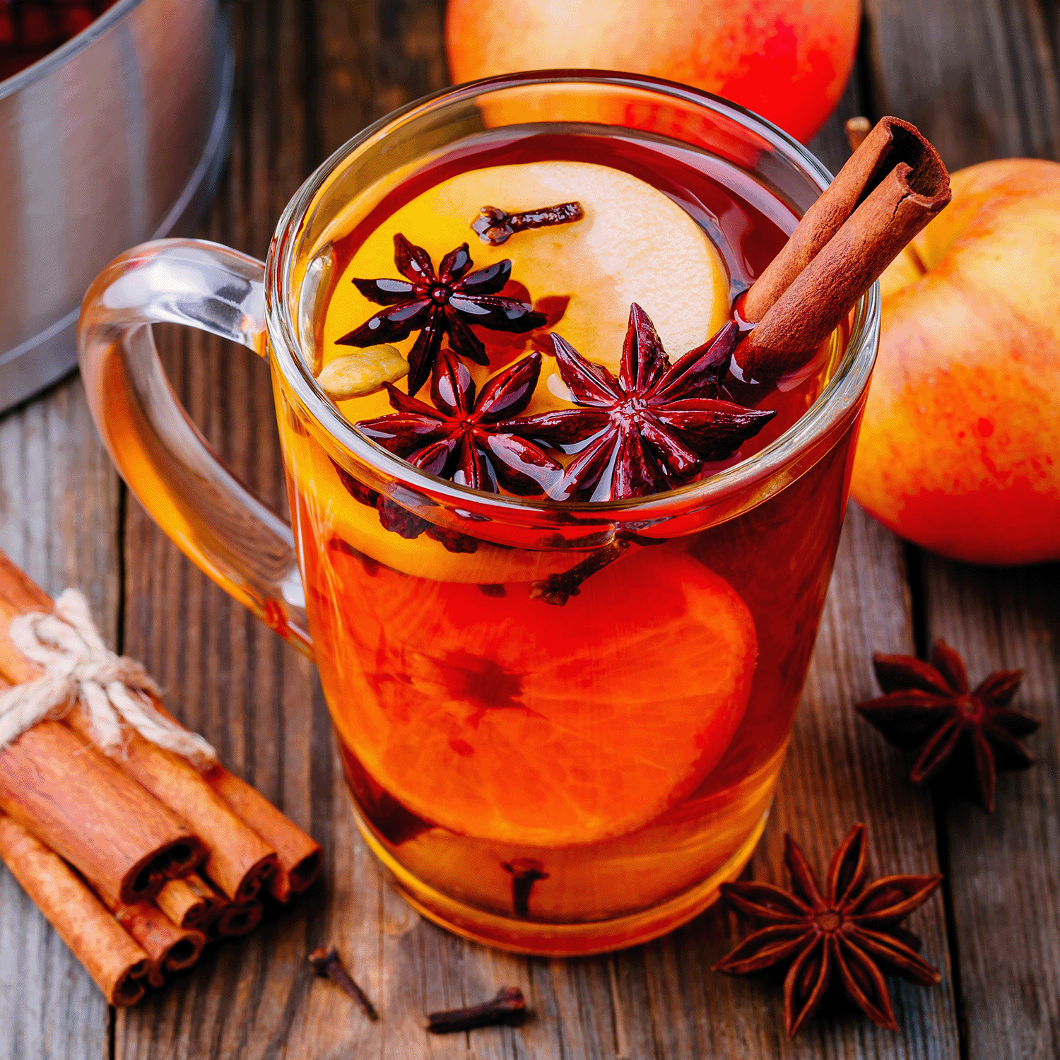 A glass mug filled with spiced apple cider, garnished with orange slices, star anise, and a cinnamon stick sits on a wooden surface. Whole apples and the Classic Apple Spice Long-Lasting Scented Jar Candle (12 oz) from the Copper Harvest Collection by Tuscany Candle® SEASONAL are scattered around.