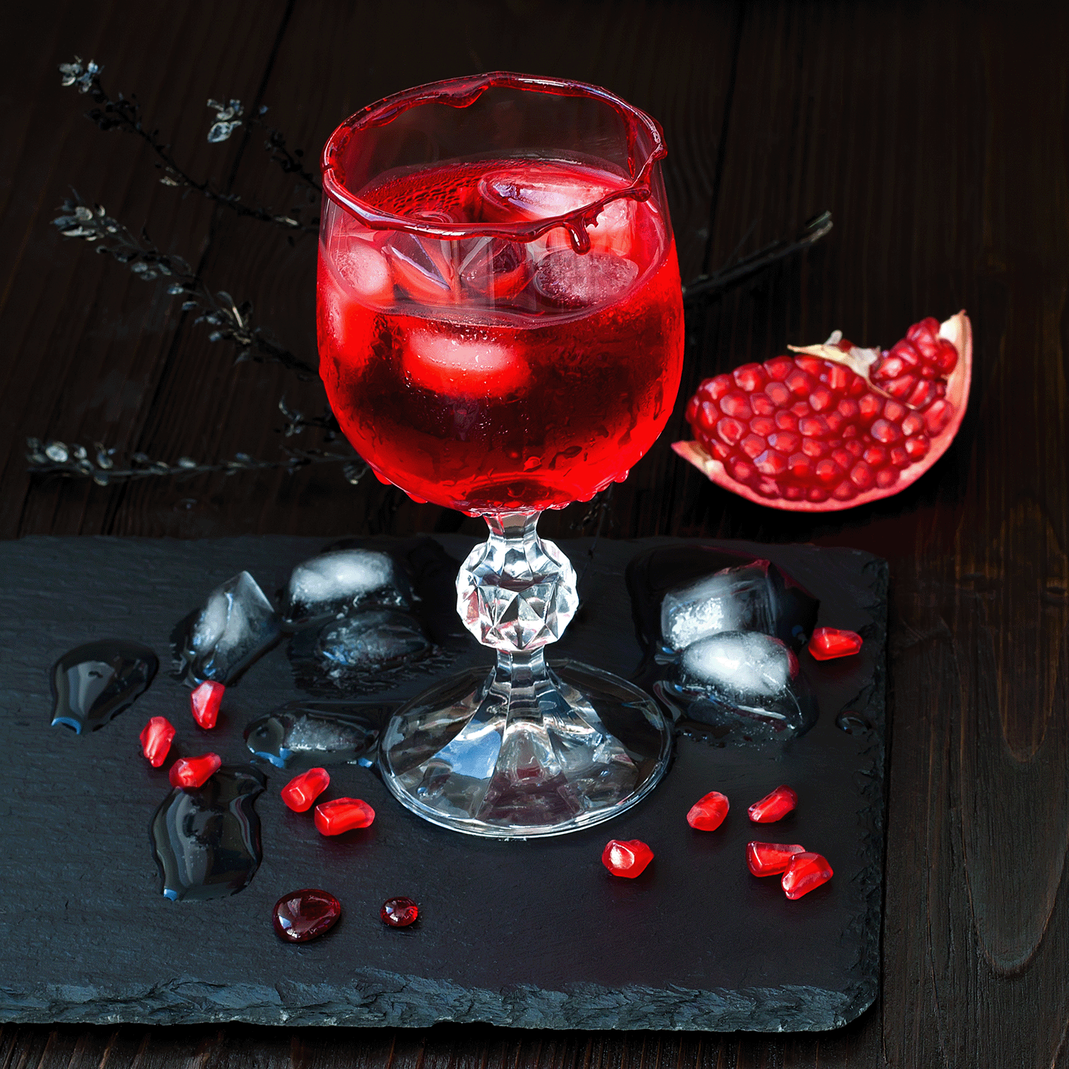 A glass of dark plum nectar with ice sits on a black stone surface, surrounded by ice cubes and scattered pomegranate seeds. Off to the side, an 18 oz jar of Tuscany Candle® SEASONAL's Bewitched Brew Long-Lasting Scented Candle adds a magical touch, while a halved pomegranate rests in the background.