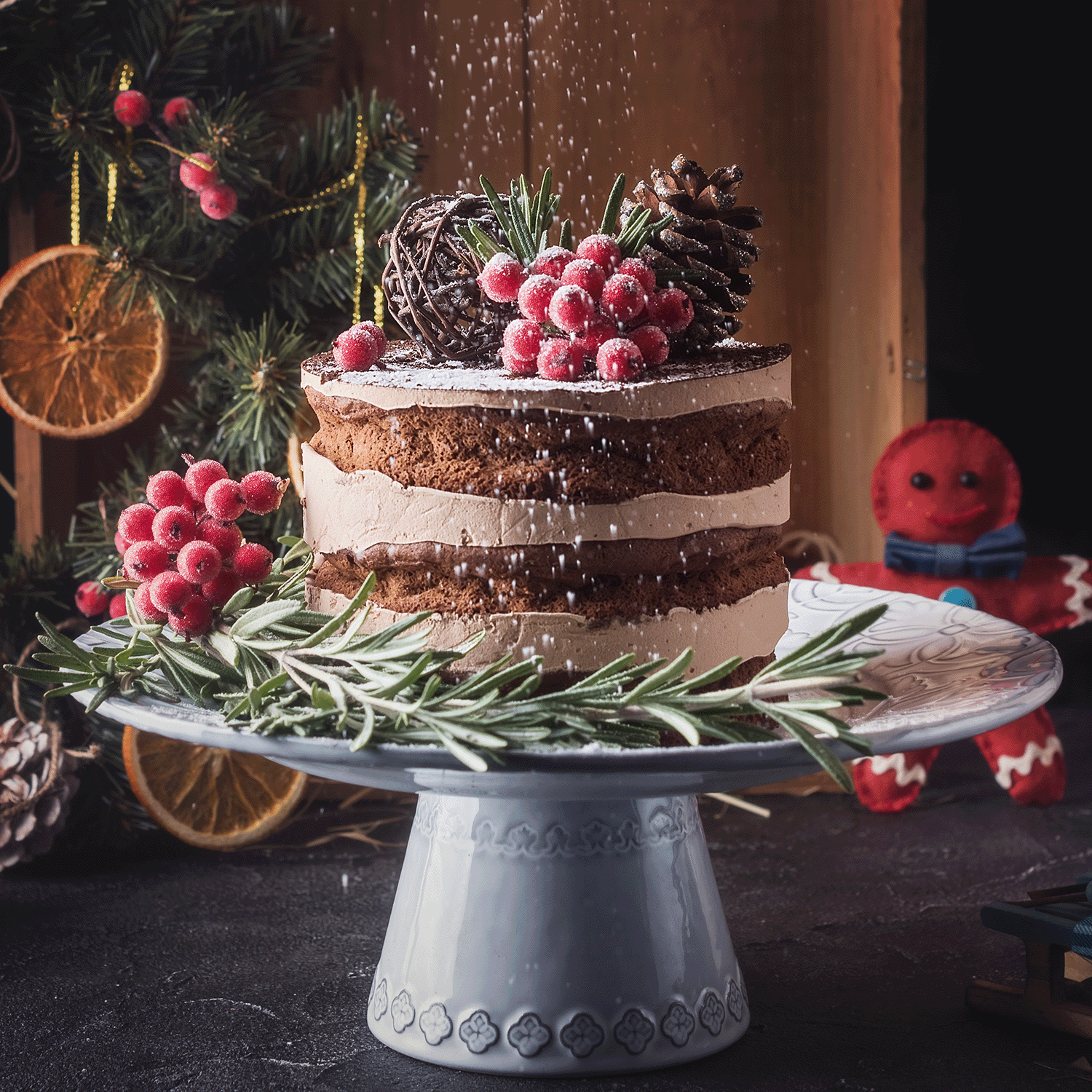 A decorated cake with chocolate layers and cream frosting rests on a white stand, adorned with red berries, pinecones, and rosemary, lightly dusted with sugar. The charming scene is completed by the festive background enhanced by the holiday fragrance of the Mulberry Bliss Long-Lasting Scented Jar Candle (18 oz) from Tuscany Candle® SEASONAL.