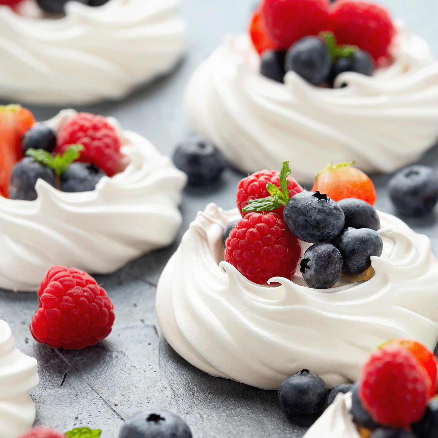 Mini pavlovas with a hint of mint are elegantly garnished with raspberries, blueberries, and fresh mint leaves on a gray surface.