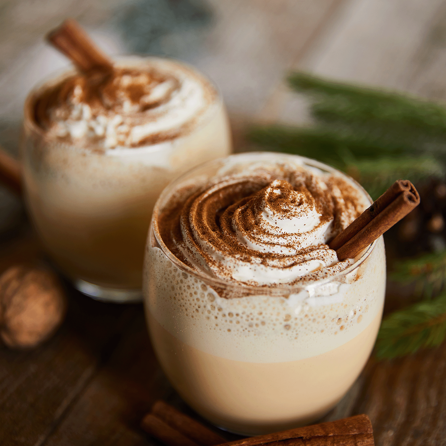 Two glasses of creamy holiday drinks topped with whipped cream, cinnamon sticks, and ground cinnamon sit alongside the Tuscany Candle® SEASONAL Gingerbread Cookies Long-Lasting Scented Jar Candle (18 oz) on a wooden table, surrounded by festive greenery in the background. The aroma evokes memories of gingerbread cookies fresh from the oven.