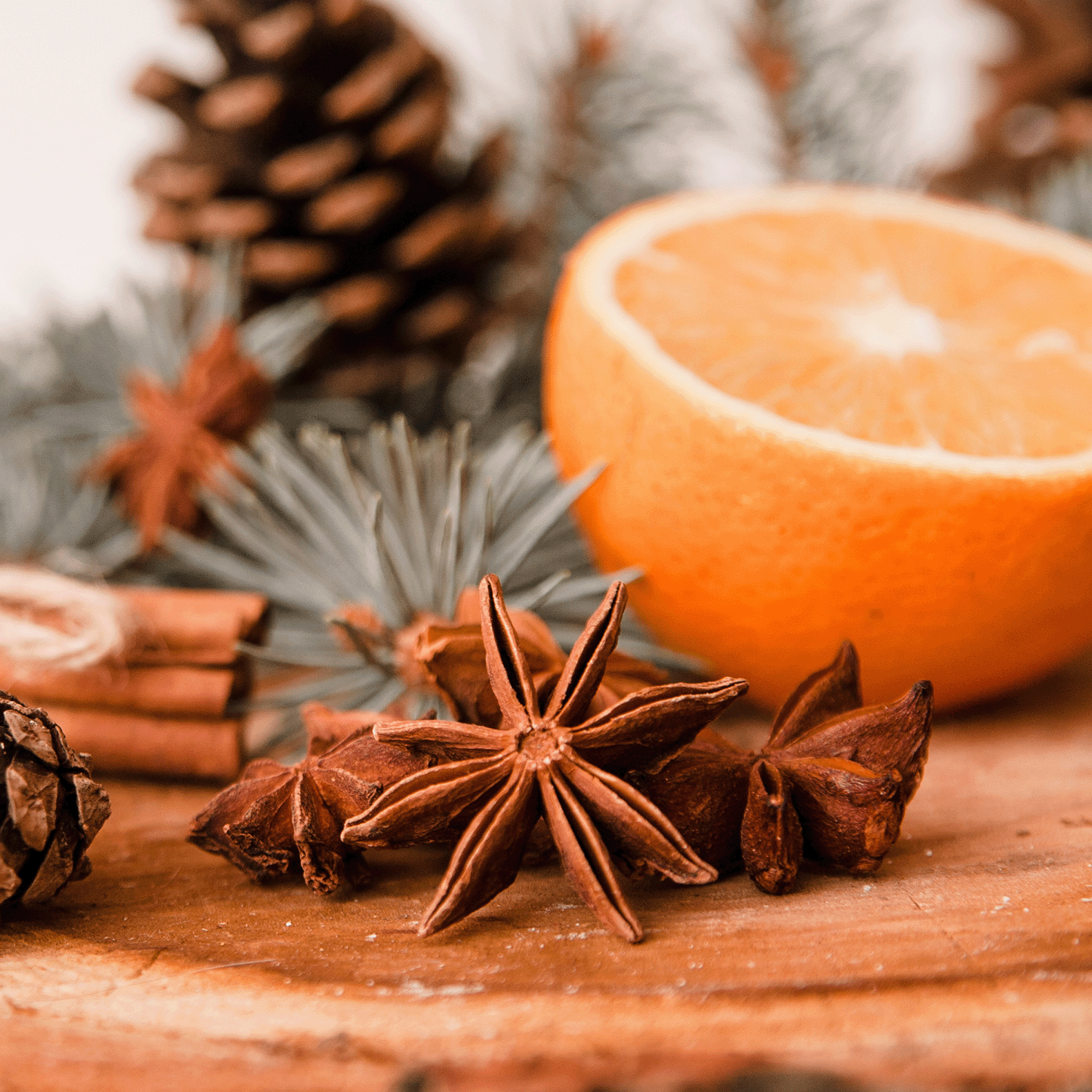 A close-up of star anise, half an orange, and a pine cone on a wooden surface is beautifully complemented by the warm aromas of the Tuscany Candle® SEASONAL Cinnamon & Spice Long-Lasting Scented Jar Candle (14 oz), set amidst pine needles and cinnamon sticks in the background.