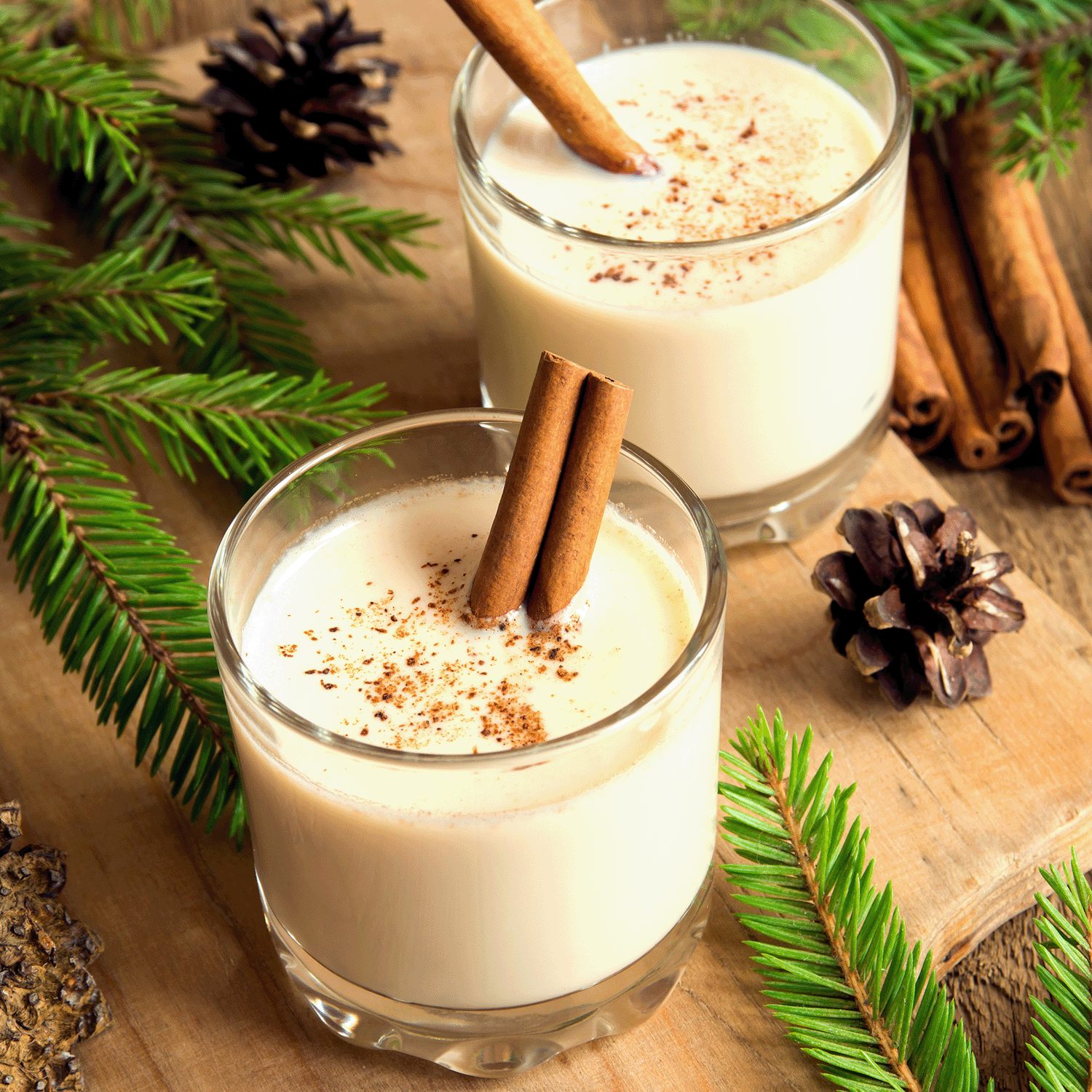 Two glasses of eggnog with cinnamon sticks, sprinkled with nutmeg, surrounded by pine branches and cones on a wooden surface evoke the essence captured by the Tuscany Candle® SEASONAL Cinnamon & Spice Long-Lasting Scented Jar Candle (14 oz), filling the air as if lit by this delightful candle that whispers warming notes of cinnamon and spice.