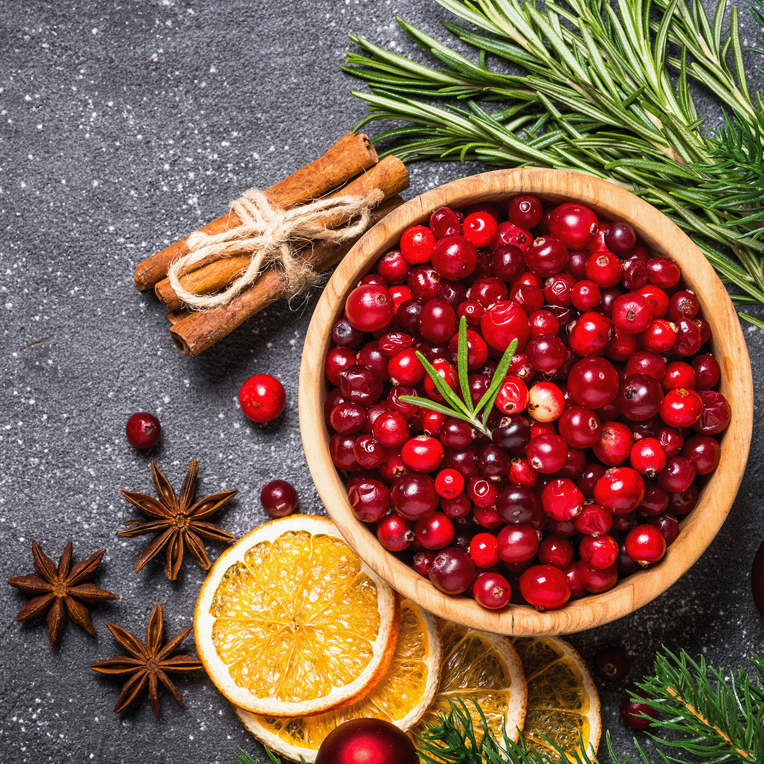 A bowl of red cranberries on a dark surface, surrounded by orange slices, star anise, cinnamon sticks, and rosemary sprigs, evokes a holiday fragrance reminiscent of the Tuscany Candle® SEASONAL Christmas Cranberry Long-Lasting Scented Jar Candle (14 oz).