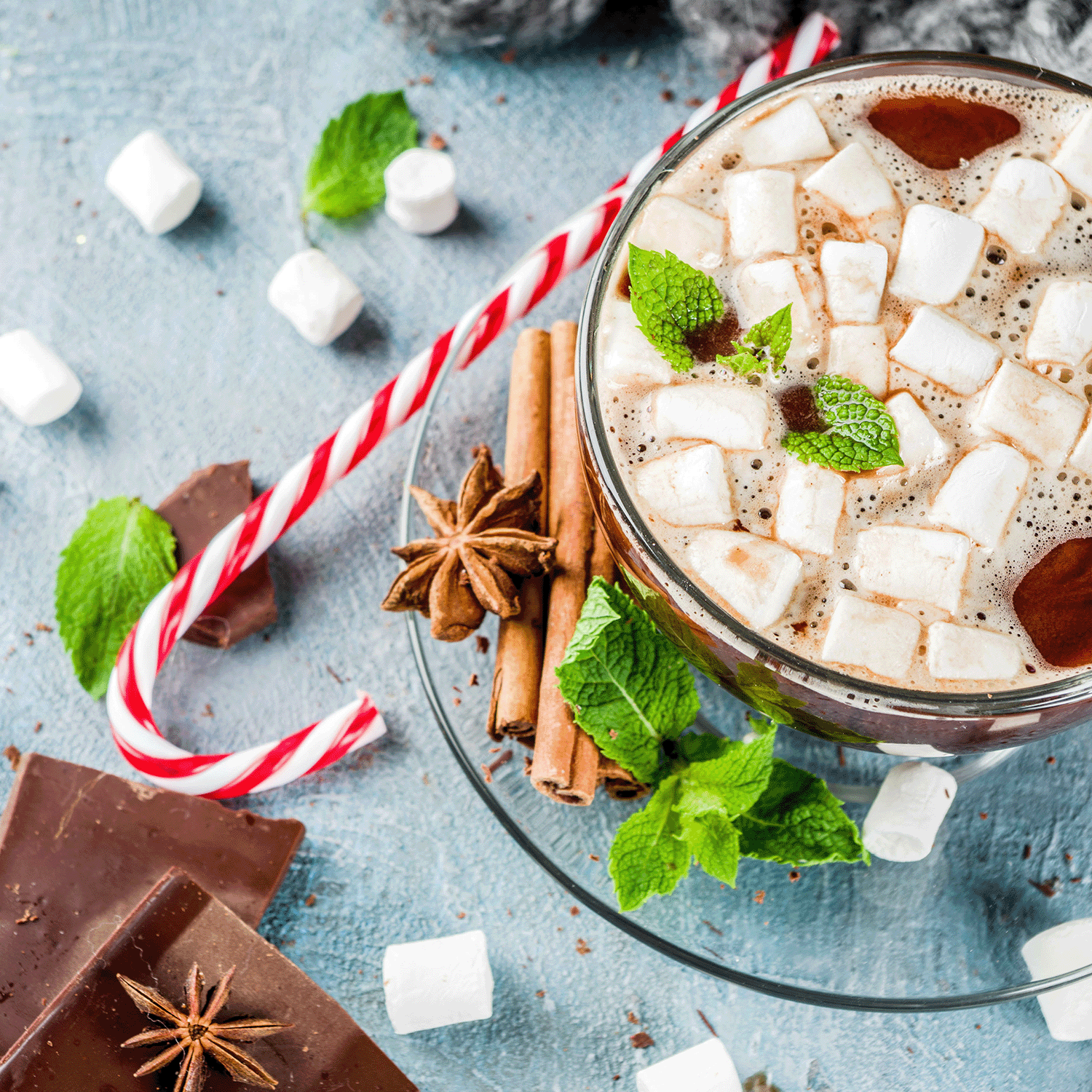 In a Winter Wonderland, a Snow Kissed Eucalyptus Long-Lasting Scented Jar Candle by Tuscany Candle® SEASONAL sits surrounded by pieces of chocolate, star anise, cinnamon sticks, a candy cane, and marshmallows on a textured surface.