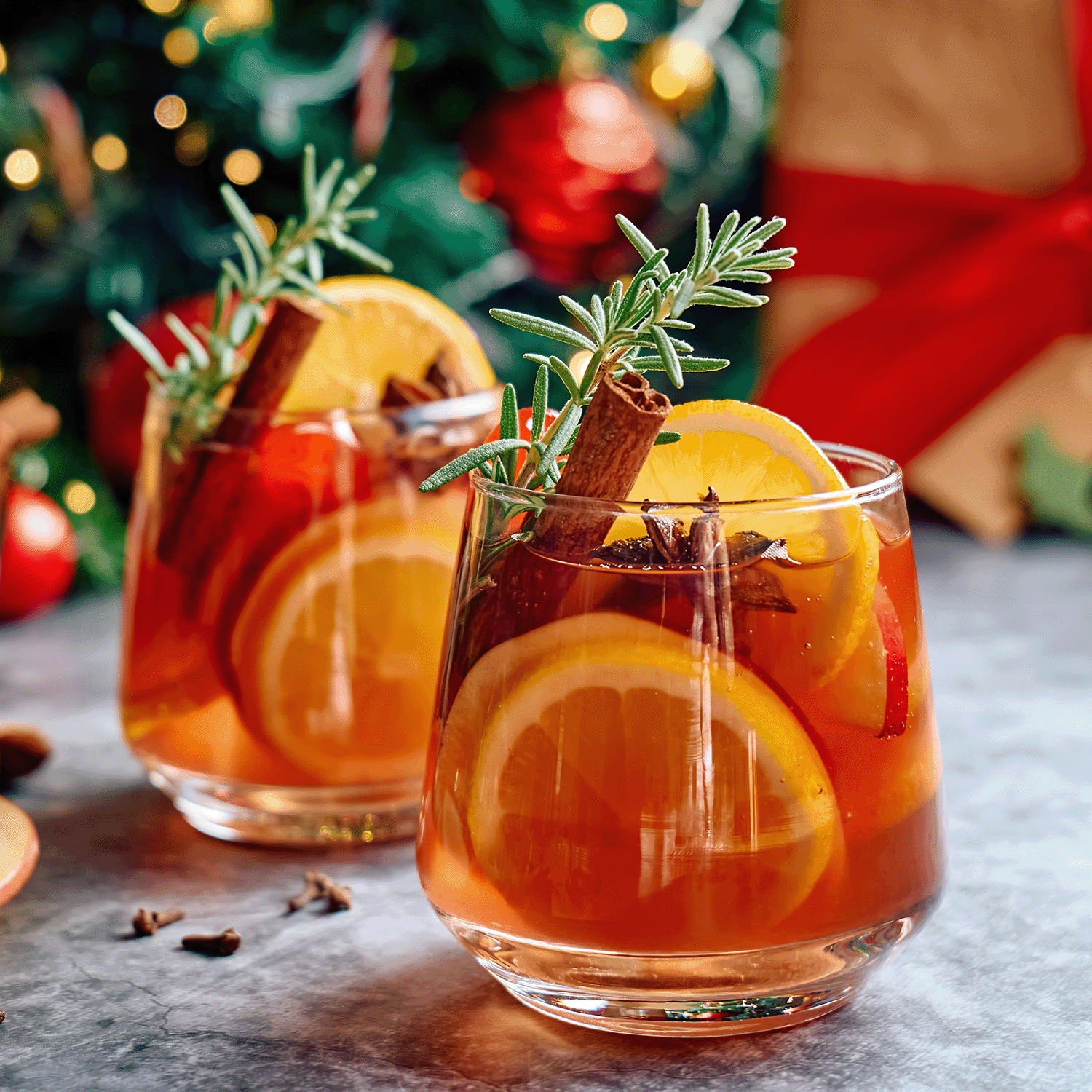 Two glasses of mulled wine adorned with orange slices, cinnamon sticks, and rosemary rest on a table. Nearby, a Tuscany Candle® SEASONAL Sparkling Sangria Long-Lasting Scented Jar Candle (14 oz) fills the air with its delightful fragrance, adding to the holiday atmosphere against a festive backdrop.