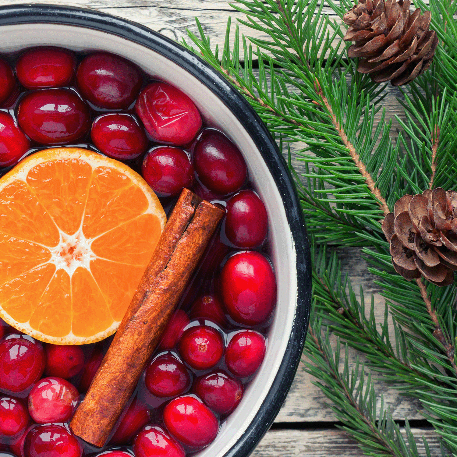 A bowl of cranberries with a halved orange and cinnamon sticks rests next to pine branches and pine cones on a wooden surface, evoking the aroma of Tuscany Candle® SEASONAL's Christmas Cranberry Scented Wax Melt (2.5 oz).