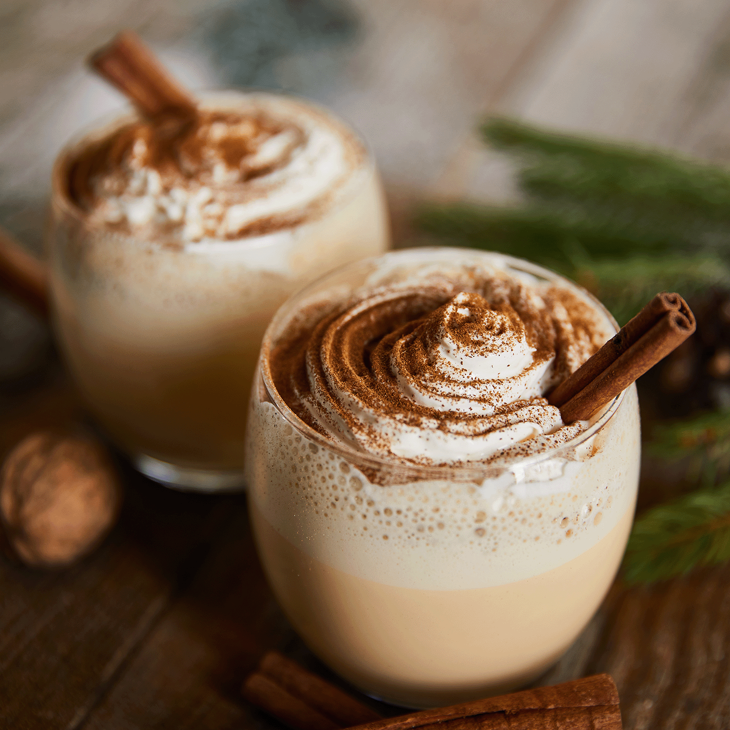 Two glasses of eggnog topped with whipped cream and cinnamon sticks sit on a wooden table, accompanied by the warm aroma of Gingerbread Jingle Scented Wax Melt from Tuscany Candle® SEASONAL, surrounded by pine branches and walnuts.