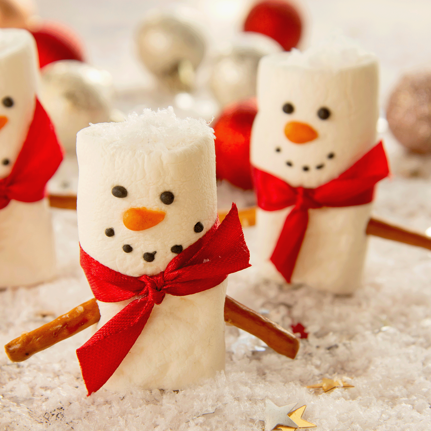 Three marshmallow snowmen featuring red scarves and pretzel stick arms are adorned with candy carrot noses, buttons, and eyes, placed on faux snow with ornaments softly blurred in the background. The scene captures the cozy essence of Tuscany Candle® SEASONAL's Hot Cocoa Nights Scented Wax Melt (2.5 oz).