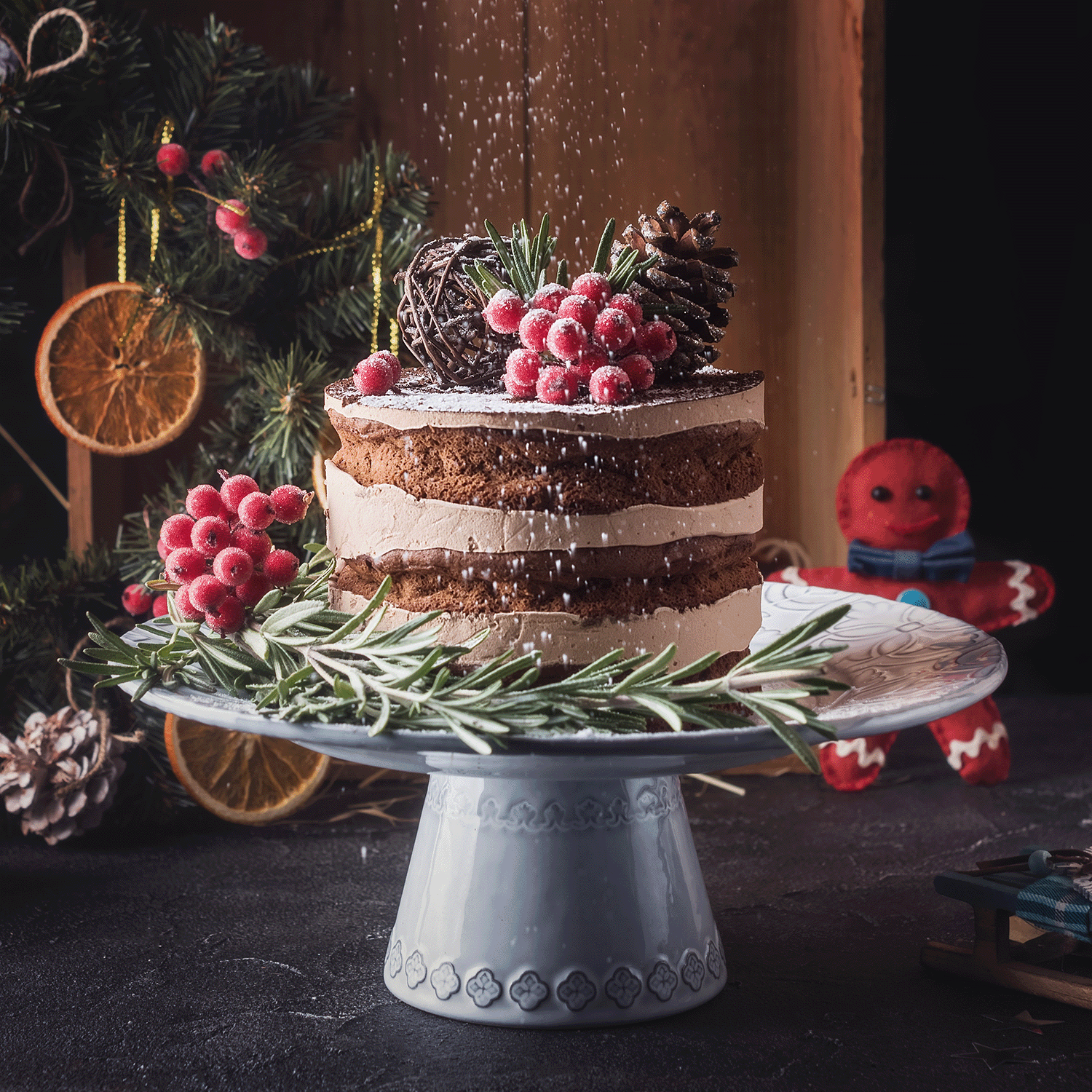 A Mulberry Bliss Scented Wax Melt from Tuscany Candle® SEASONAL, featuring a festive theme with red berries, rosemary, and a pine cone set on a white display. It is accented with dried oranges and includes a gingerbread figure in the backdrop, lightly dusted with powdered sugar.