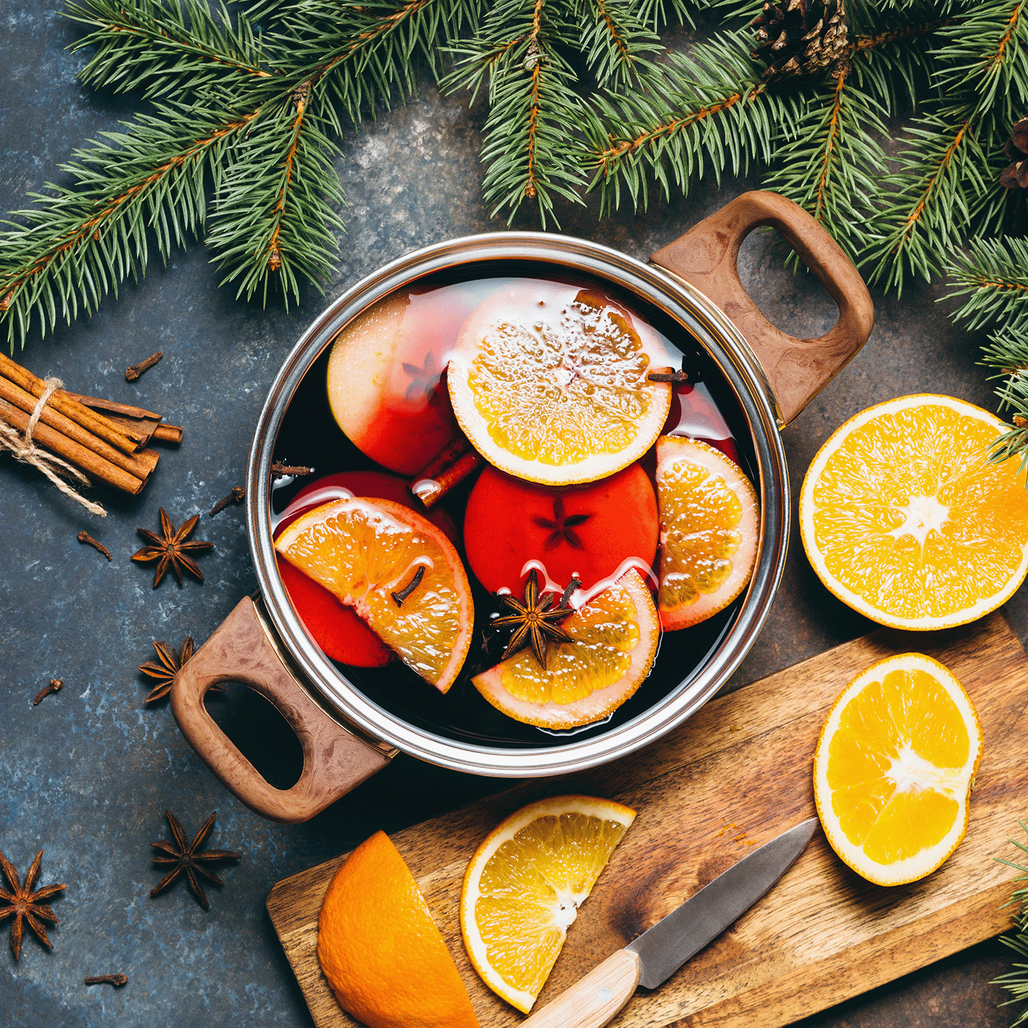 A pot of mulled wine infused with sliced apples, oranges, and spices creates a cozy scene. To the side, a cutting board holds neatly sliced oranges alongside a knife, all encircled by pine branches. Add to the ambiance with Tuscany Candle® SEASONAL's Sparkling Sangria Scented Wax Melt (2.5 oz) for an inviting fragrance.