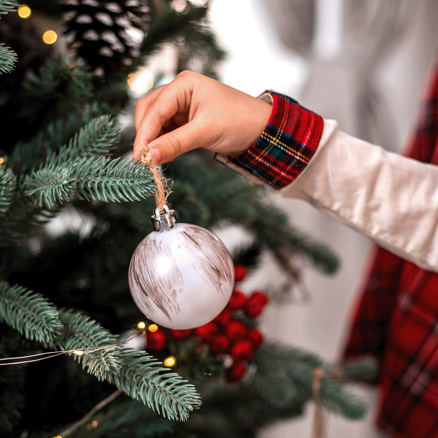 A hand in a plaid-accented sleeve places the Tree Trimming Scented Wax Melt by Tuscany Candle® SEASONAL on a decorated Christmas tree adorned with pine cones and red berries.