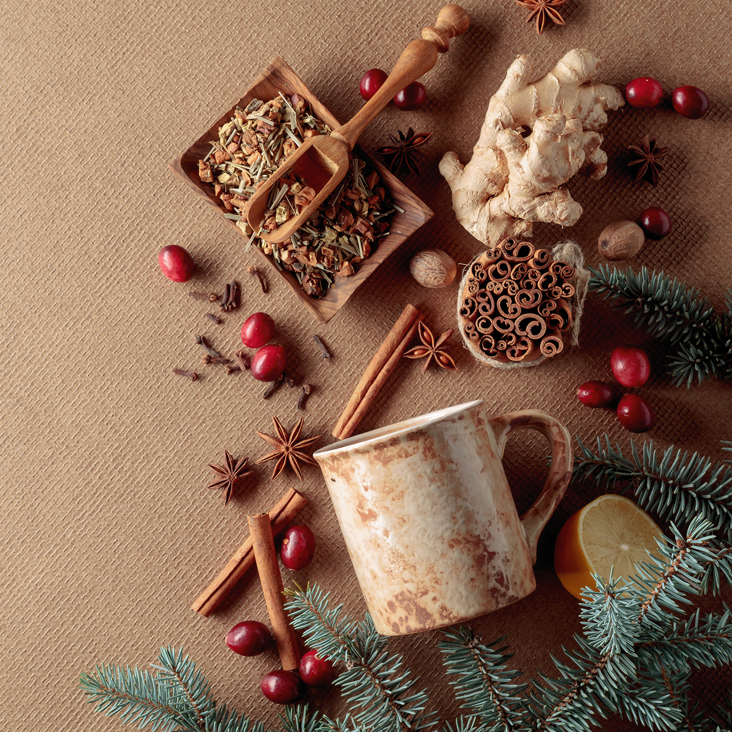 A rustic mug rests on a textured surface, surrounded by loose tea leaves, spices, ginger, cranberries, pine branches, and a lemon slice. The Good Tidings of Cinnamon & Joy Scented Jar Candle (12 oz) from Tuscany Candle® SEASONAL adds warmth to this cozy and festive arrangement, perfect for enhancing your holiday decor.