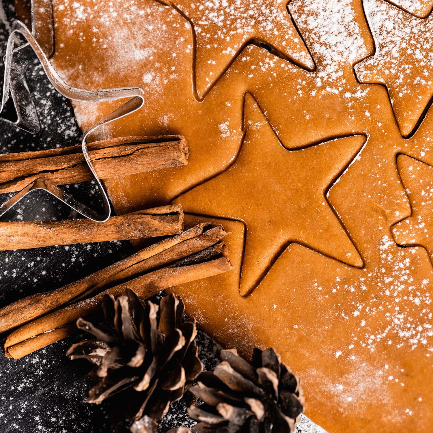 The Season's Greetings Scented Jar Candle by Tuscany Candle® SEASONAL sits amid a scene of rolled dough adorned with star-shaped cookie cutters, flanked by cinnamon sticks and pinecones lightly dusted with flour, encapsulating the spirit of holiday décor.