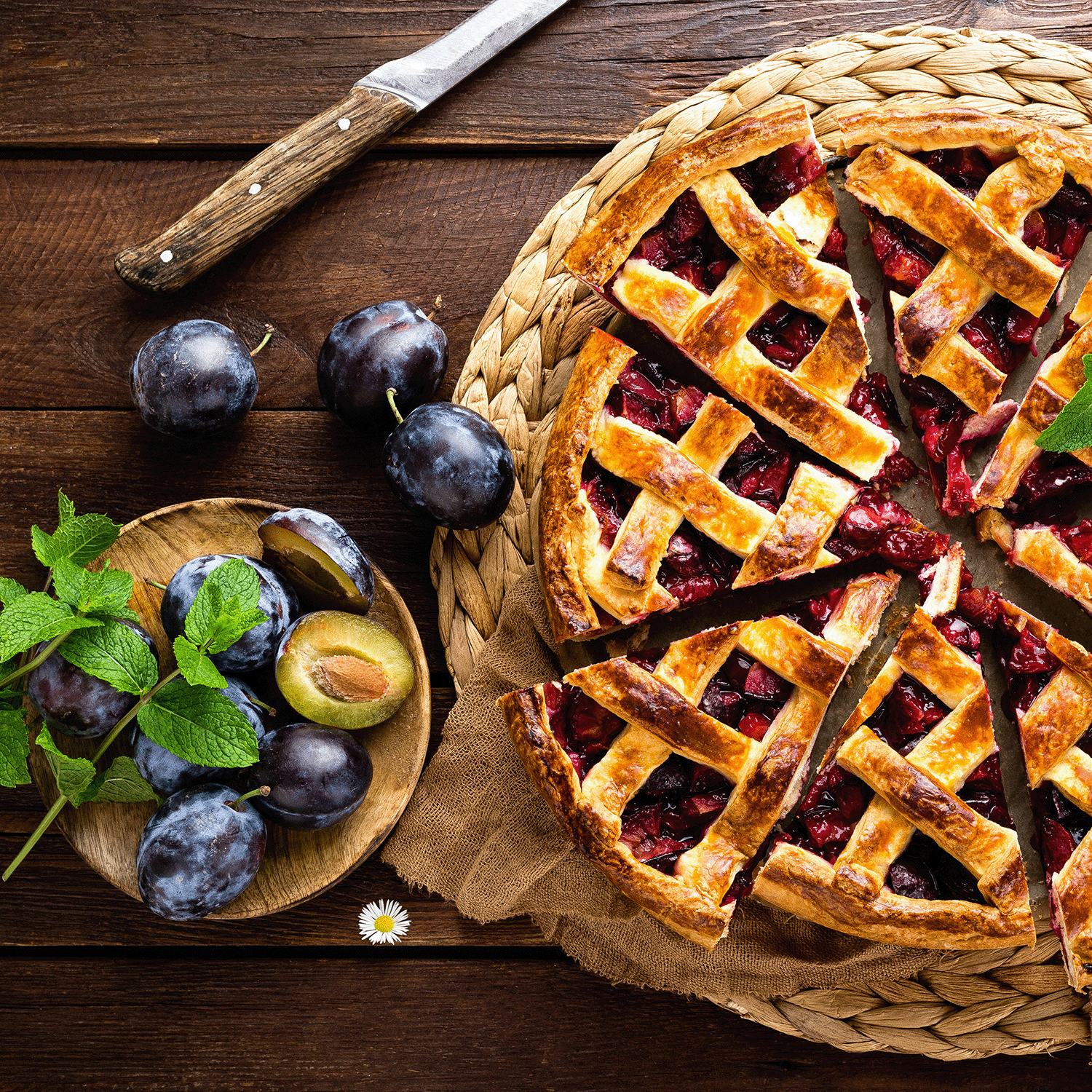 A sliced lattice-topped pie on a woven mat, its aroma complemented by a nearby 12 oz Sugar Plums Scented Jar Candle from Tuscany Candle® SEASONAL, fills the air with a delightful holiday fragrance. A small bowl of plums sits beside it on the wooden table.