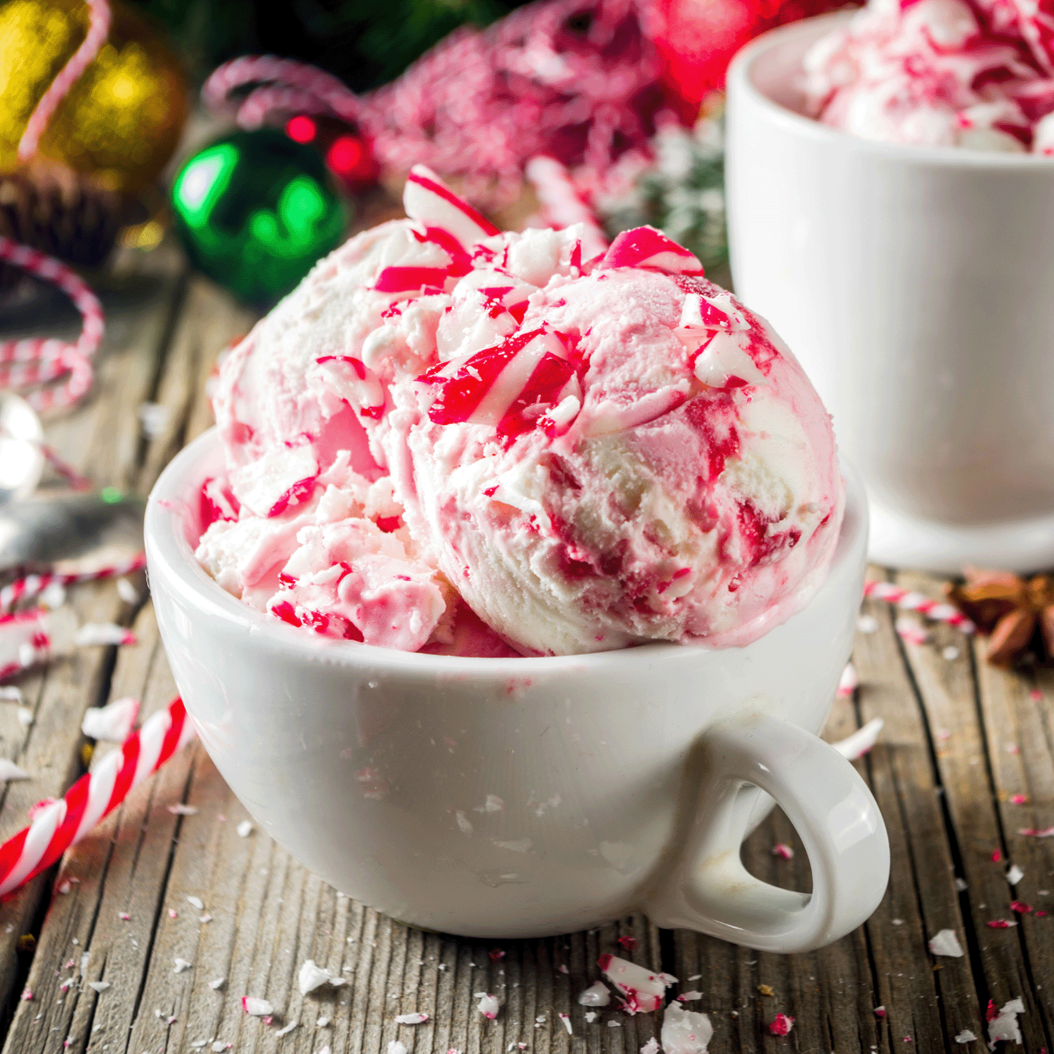 A cup of peppermint ice cream with crushed candy canes is positioned on a wooden table adorned with festive holiday décor, including the 15 oz Peppermint & Spruce Scented Jar Candle from Tuscany Candle® SEASONAL.