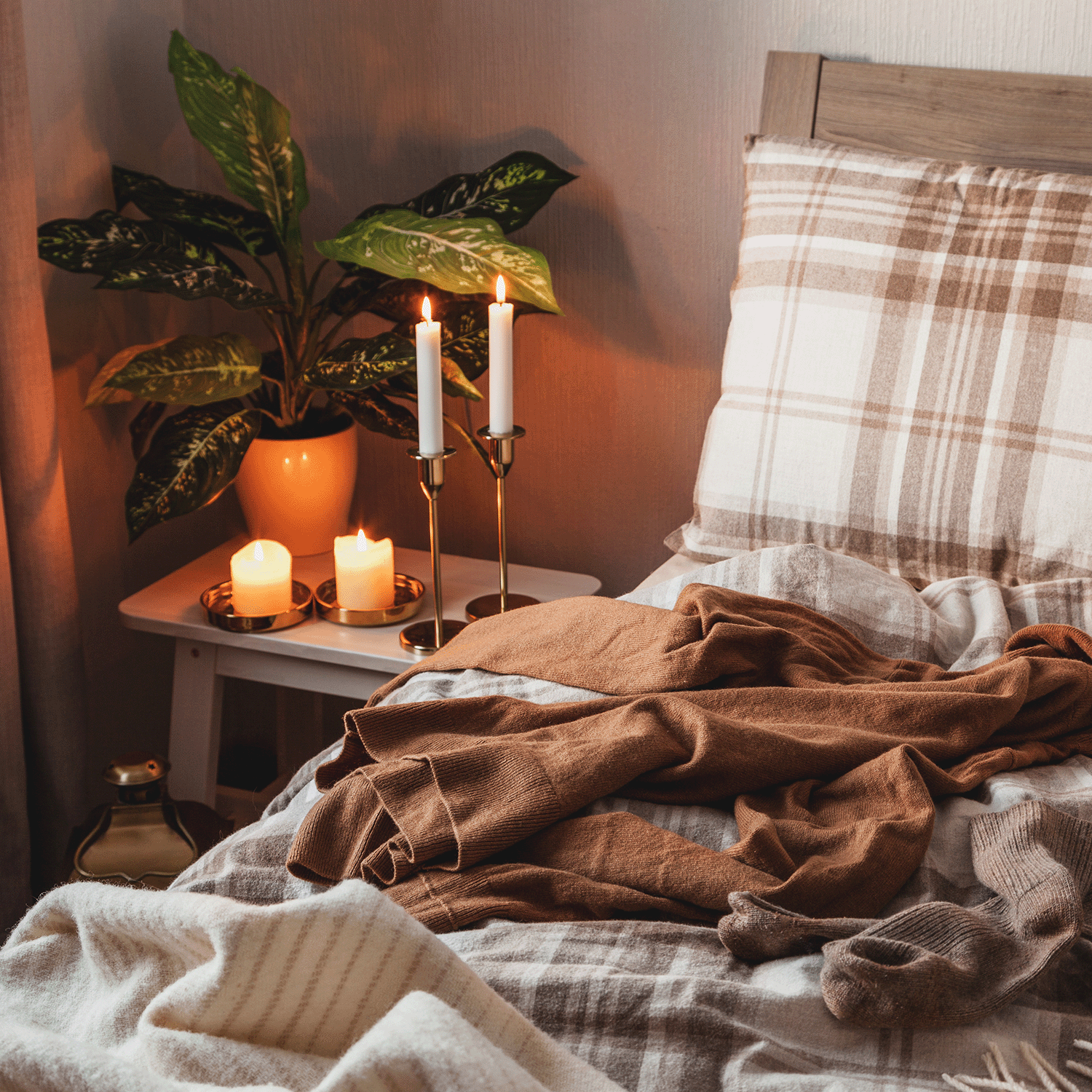 Cozy bedroom with plaid bedding, lit candles on a small table, and a potted plant. A brown sweater and socks are casually draped on the bed. The gentle glow from the Cozy Autumn Flannels Long-Lasting Scented Jar Candle (18 oz) by Tuscany Candle® SEASONAL, part of our limited Autumn Flannels Collection, brings an extra touch of warmth to the space.