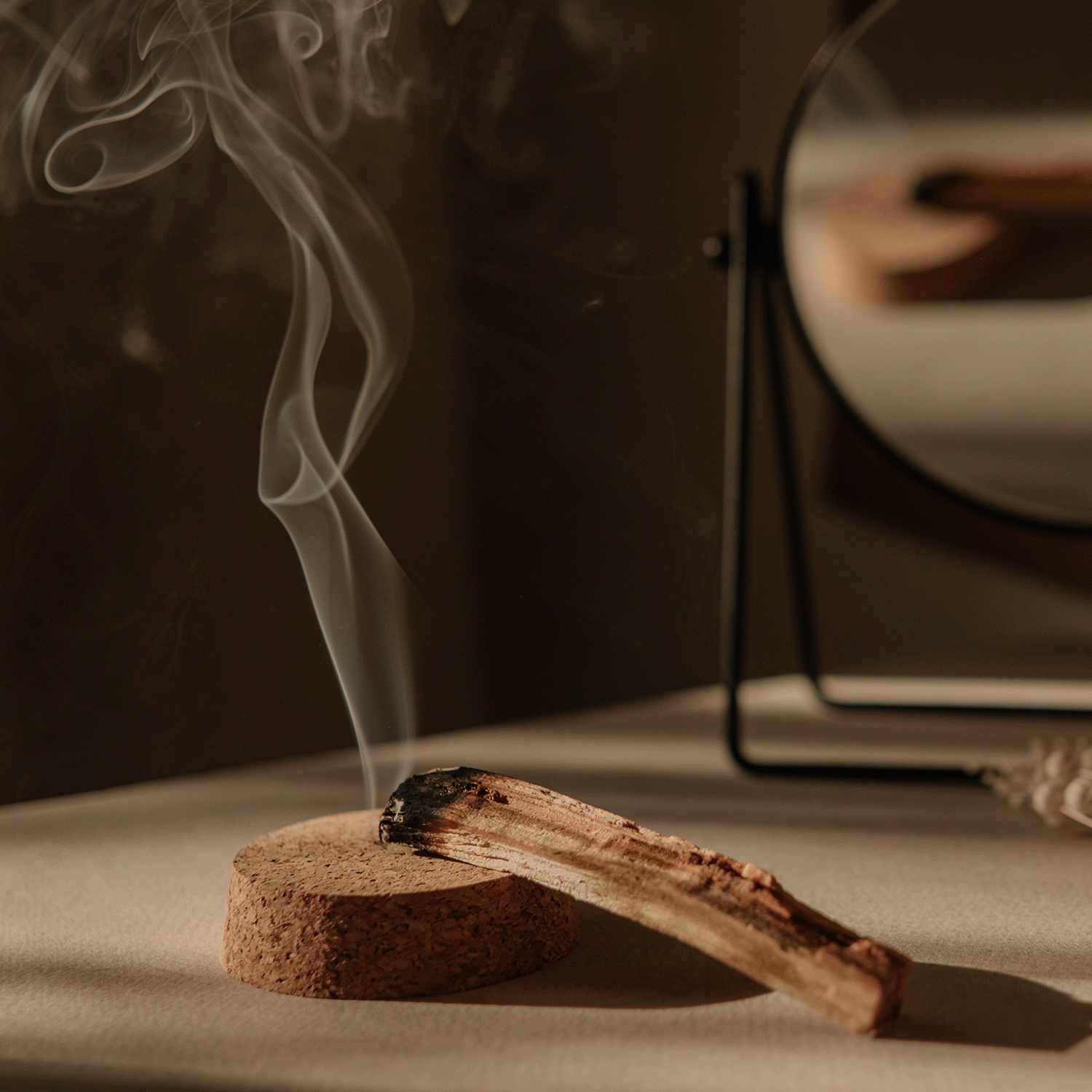 A wooden stick smoldering and releasing smoke rests on a cork base with a mirror in the background, casting shadows on a surface. Part of our Autumn Flannels Collection limited-run, this scene pairs perfectly with the Cozy Autumn Flannels Long-Lasting Scented Jar Candle (18 oz) by Tuscany Candle® SEASONAL nearby.