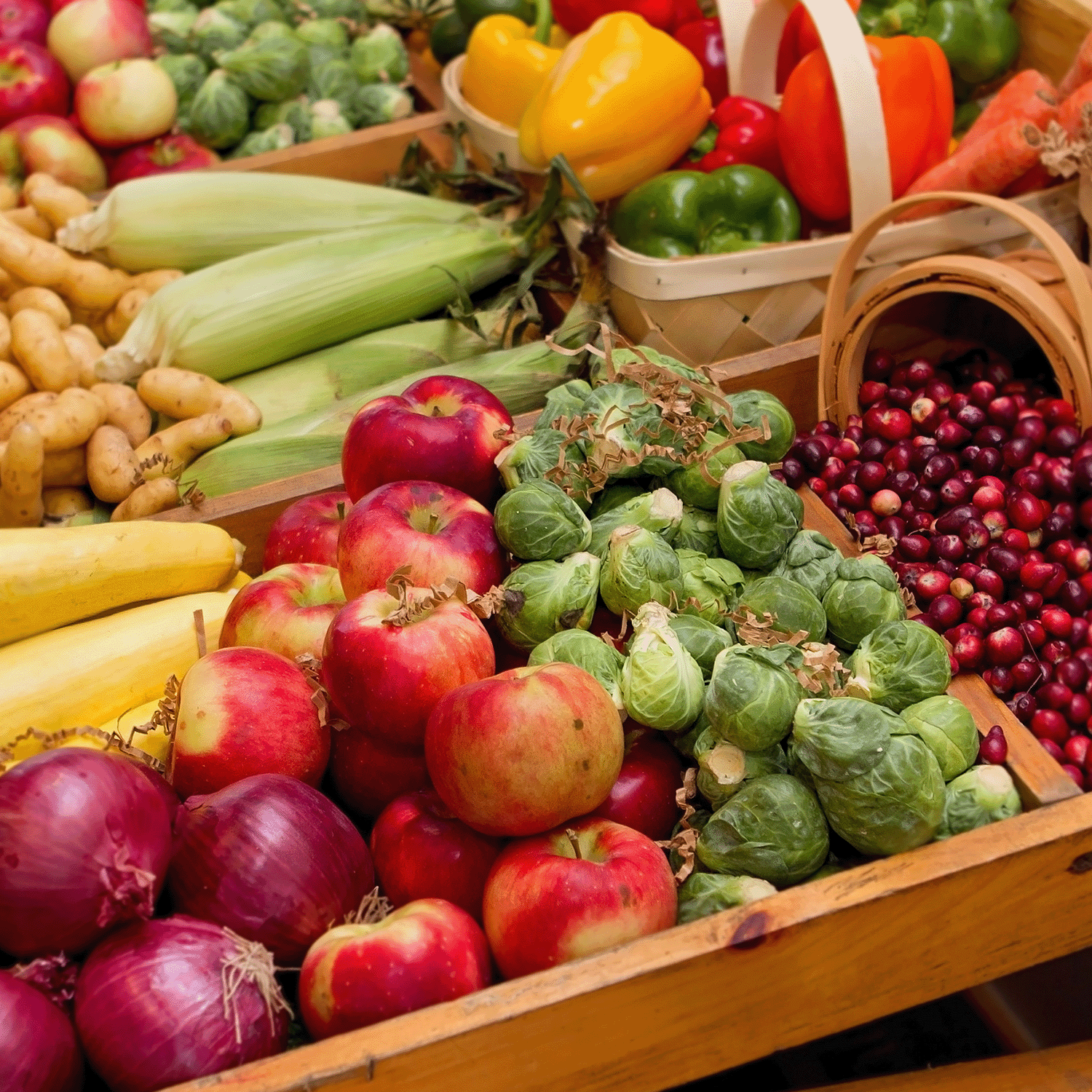 A vibrant farmers market display featuring apples, red onions, Brussels sprouts, bananas, corn, potatoes, peppers, carrots, cranberries and baskets of assorted vegetables. Among the fresh produce stands a delightful Tuscany Candle® SEASONAL Farmers Market Glazed Pistachios Long-Lasting Scented Jar Candle (18 oz) from the Autumn Flannels Collection.