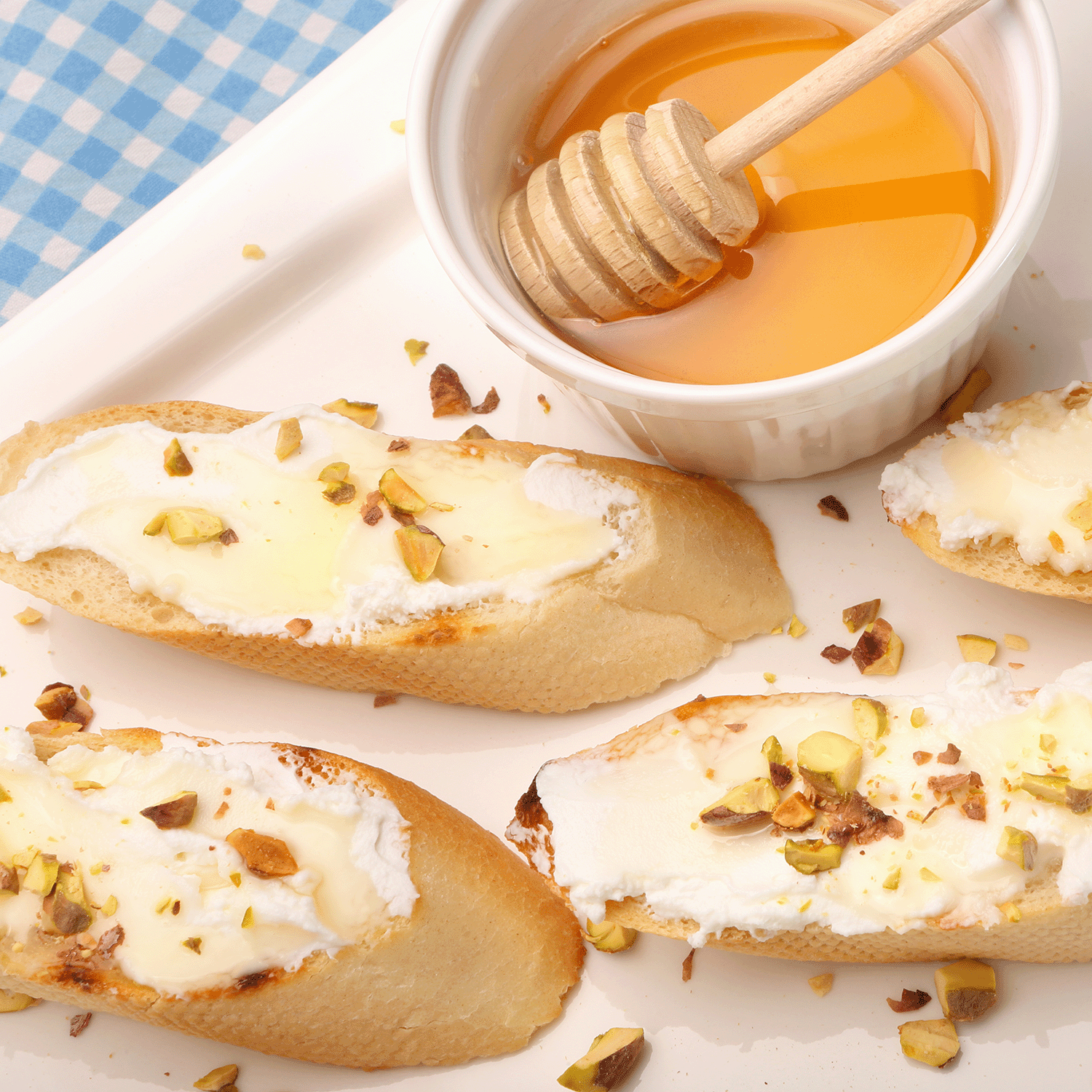 Toasted baguette slices with cream cheese and pistachios are elegantly placed beside a honey and cardamom infusion on a checkered tablecloth, alongside the Easter Morning: Cardamom & Honey Scented Easter Candle (12 oz) by Tuscany Candle® SEASONAL.