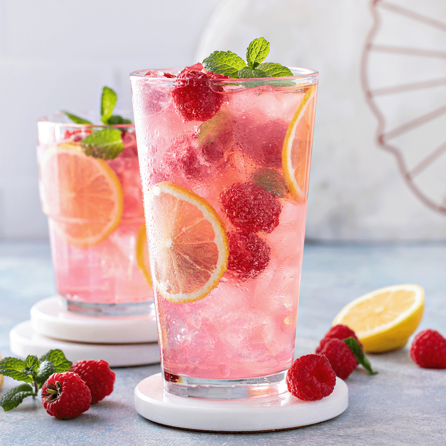 Two glasses of pink raspberry lemonade with lemon slices and mint, filled with ice, sit on white coasters. Fresh raspberries and a lemon half rest on a blue surface, evoking the sweet charm of Tuscany Candle® SEASONAL's 18 oz Cotton Tails: Raspberry Scented Easter Candle.