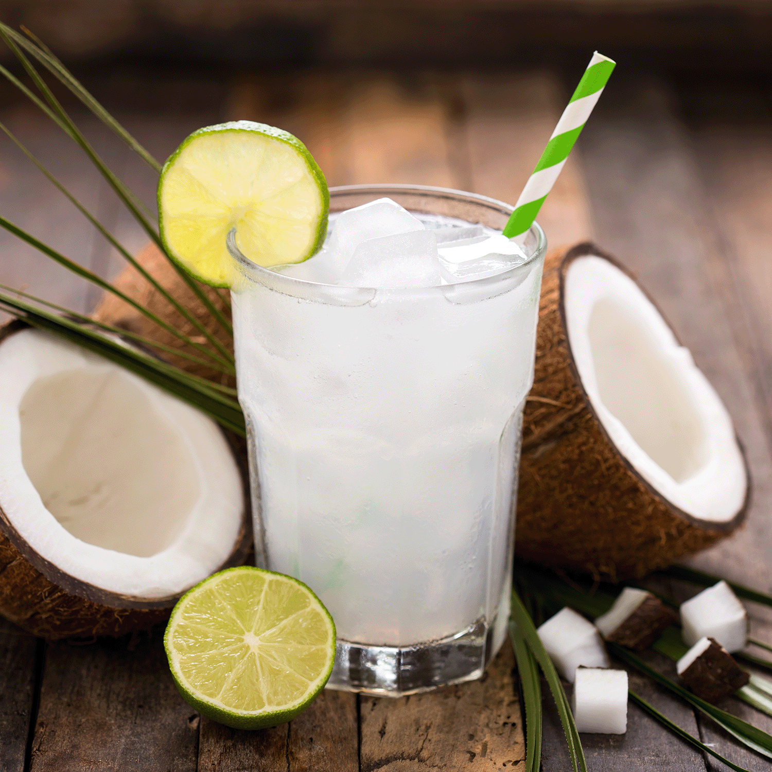 A glass of coconut water with ice, lime slices, and a green-striped straw evokes Tuscany Candle® SEASONAL's Coconut Blooms: Tropical Scented Spring Candle (18 oz), surrounded by halved coconuts and lime on wood, capturing the refreshing vibe of its tropical essence.