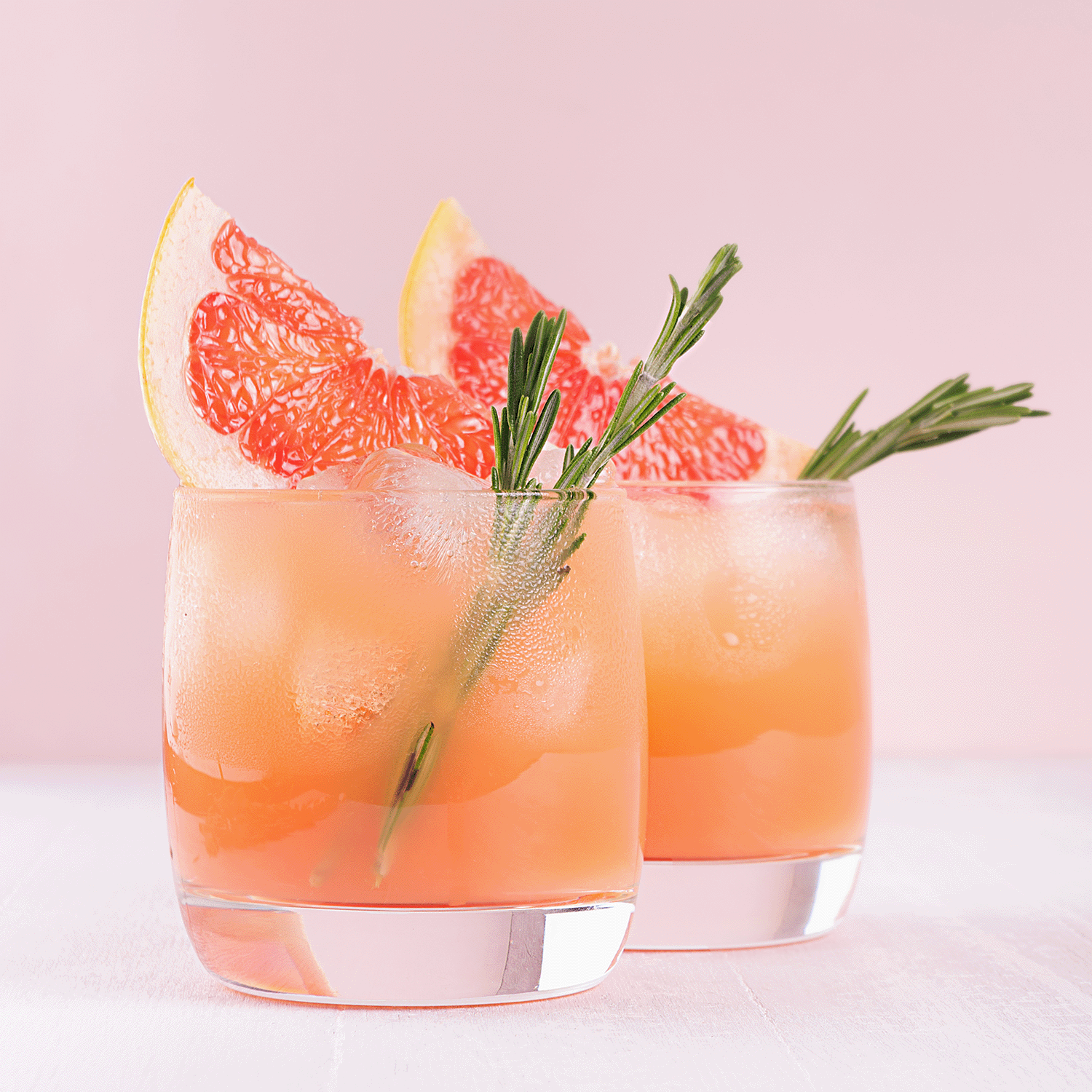 Two glasses of grapefruit cocktail with ice, garnished with rosemary sprigs and grapefruit slices, set against the soft pink background matching the Citrus Splash: Tropical Scented Spring Candle by Tuscany Candle® SEASONAL.