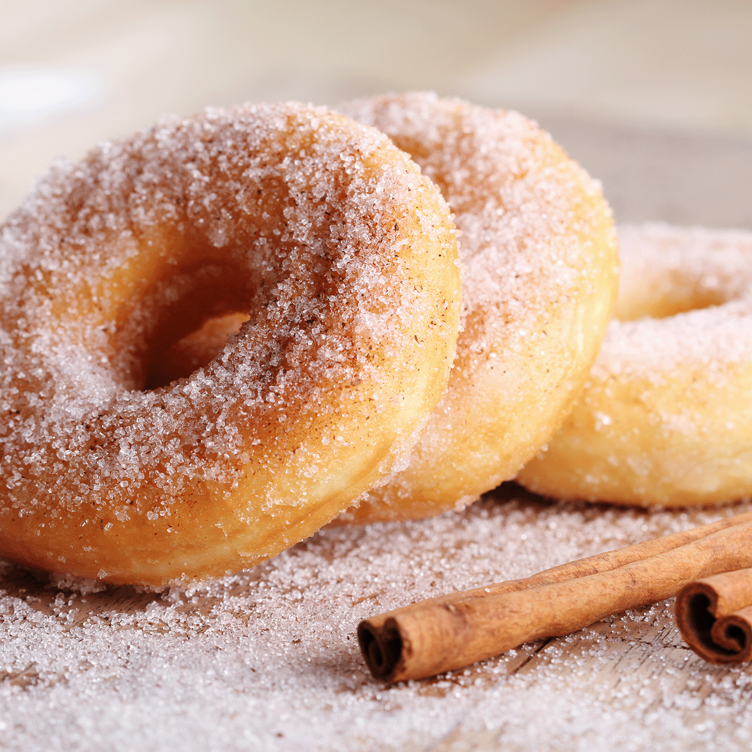 On a wooden surface sit three sugar-coated cinnamon donuts with two cinnamon sticks, reminiscent of the warm aroma of Tuscany Candle® SEASONAL's 18 oz Cinnamon Donut: Cinnamon & Sugar Scented Spring Candle.