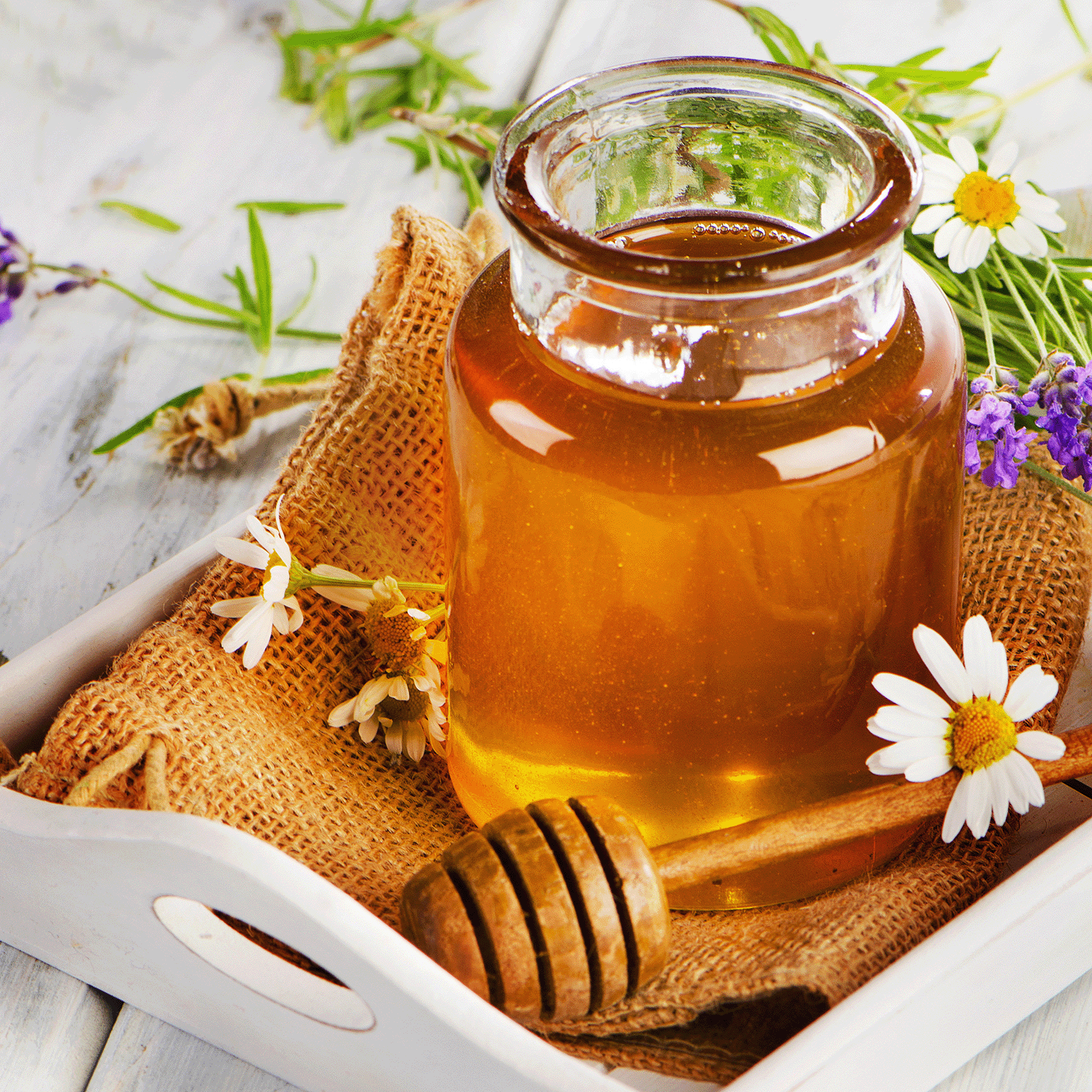 A jar of honey on burlap in a white tray, adorned with daisies, lavender, and a honey dipper evokes the soothing aroma of Tuscany Candle® SEASONAL's Iced Chai: Cardamom & Honey Scented Spring Candle (18 oz).