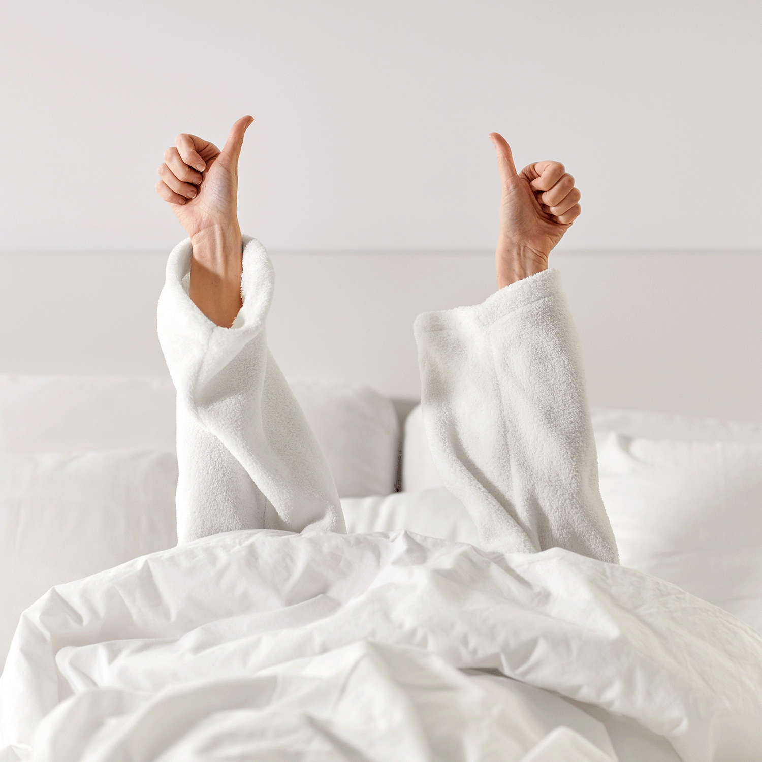 A person in a white robe lies in bed with arms raised, giving two thumbs up and surrounded by the soothing glow of Tuscany Candle® SEASONAL's Lime Blossom: Linen Scented Spring Candle (18 oz).