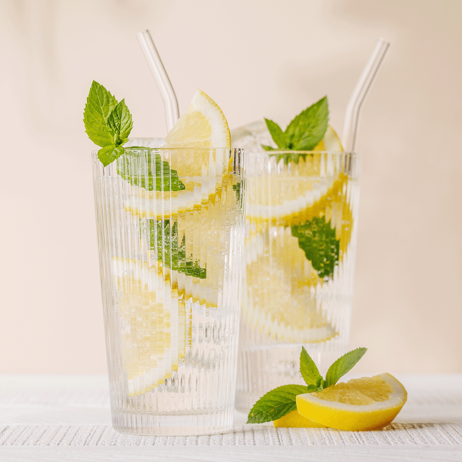 Two clear glasses filled with lemon slices, mint leaves, ice cubes, and straws capture the essence of Tuscany Candle® SEASONAL's refreshing Orange Mojito: Bergamot & Yuzu Scented Spring Candle (14 oz).