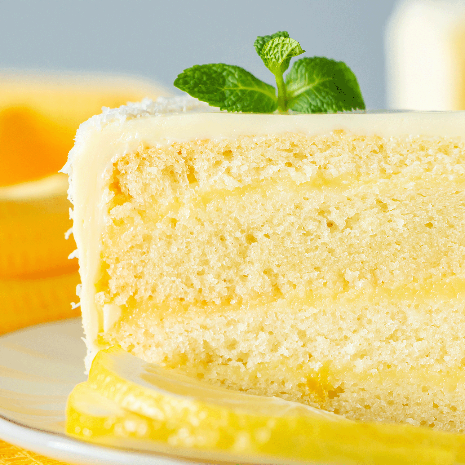 A slice of frosted lemon cake with a mint sprig rests on a white plate against a yellow fabric backdrop, evoking a cozy afternoon while the Tuscany Candle® SEASONAL Vanilla Lemonade: Meyer Lemon Scented Spring Candle (14 oz) fills the air with sweet and citrusy aromas.