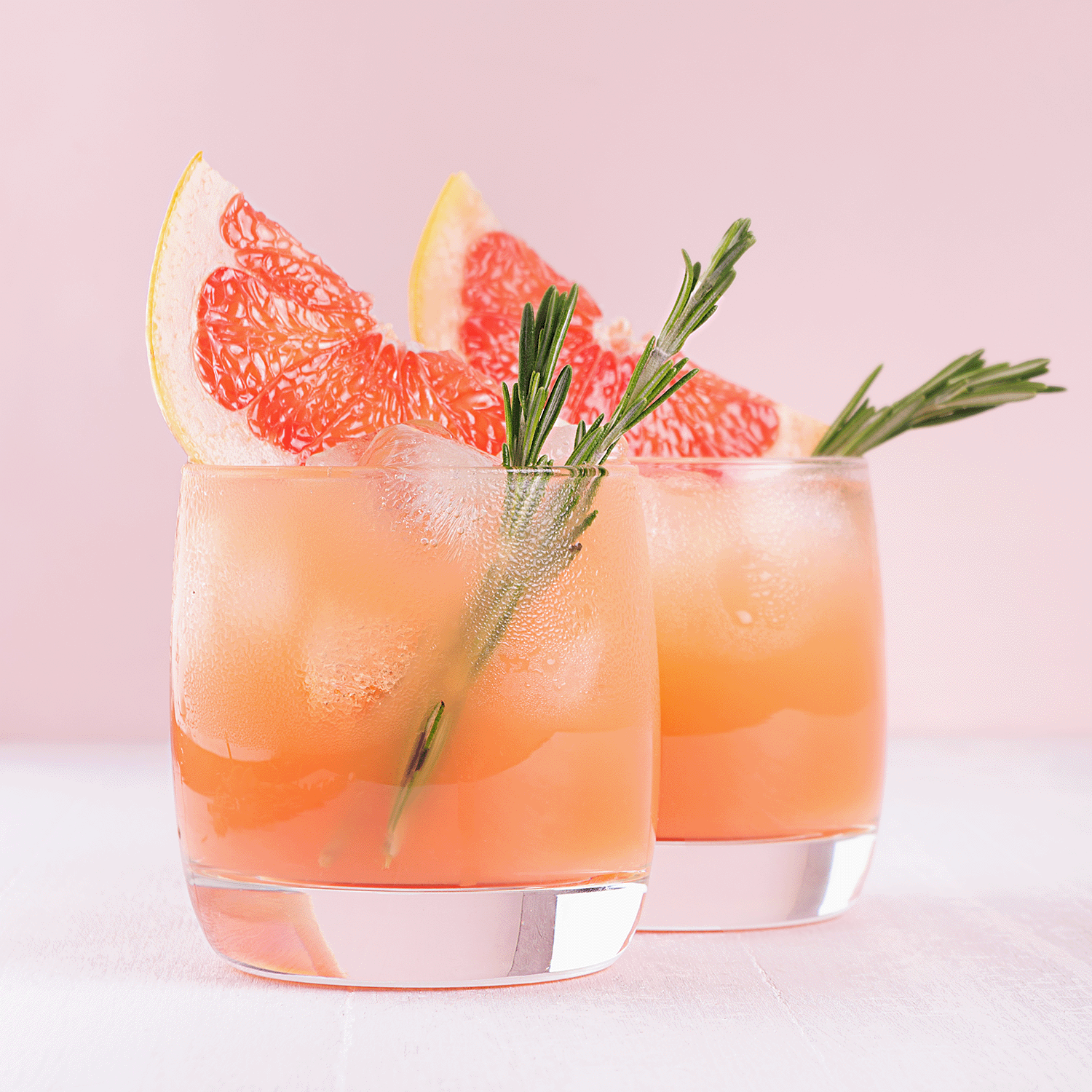 Two glasses containing pink grapefruit cocktails, each infused with a Citrus Splash, decorated with wedges of grapefruit and rosemary sprigs, placed against a pastel pink backdrop.