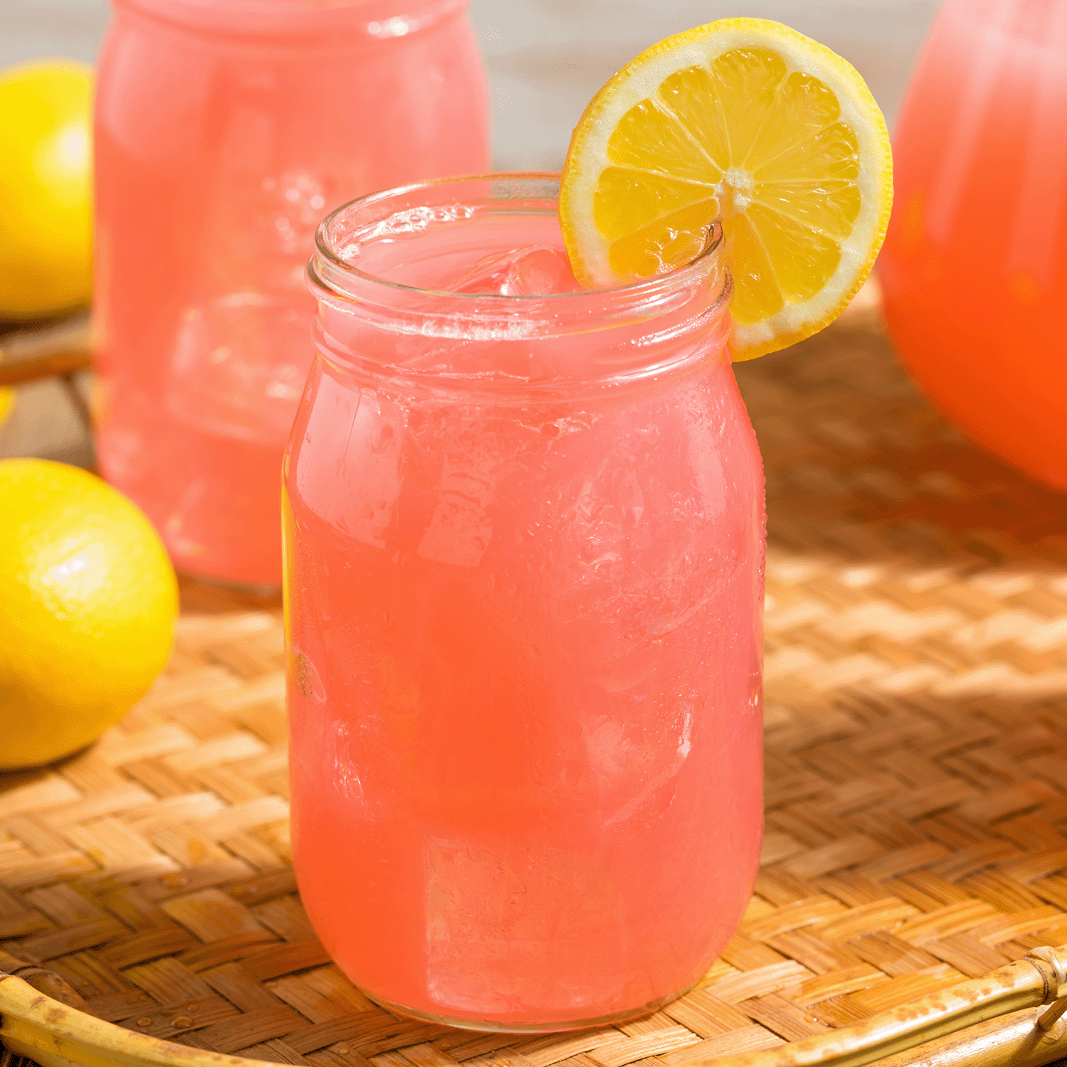 A 12 oz Frosé Lemonade: Tart Citrus Scented Candle from Tuscany Candle® SEASONAL sits on a woven tray, evoking the look of pink lemonade with ice and lemon, with whole lemons in the background as part of the Gourmet Sorbet Collection.