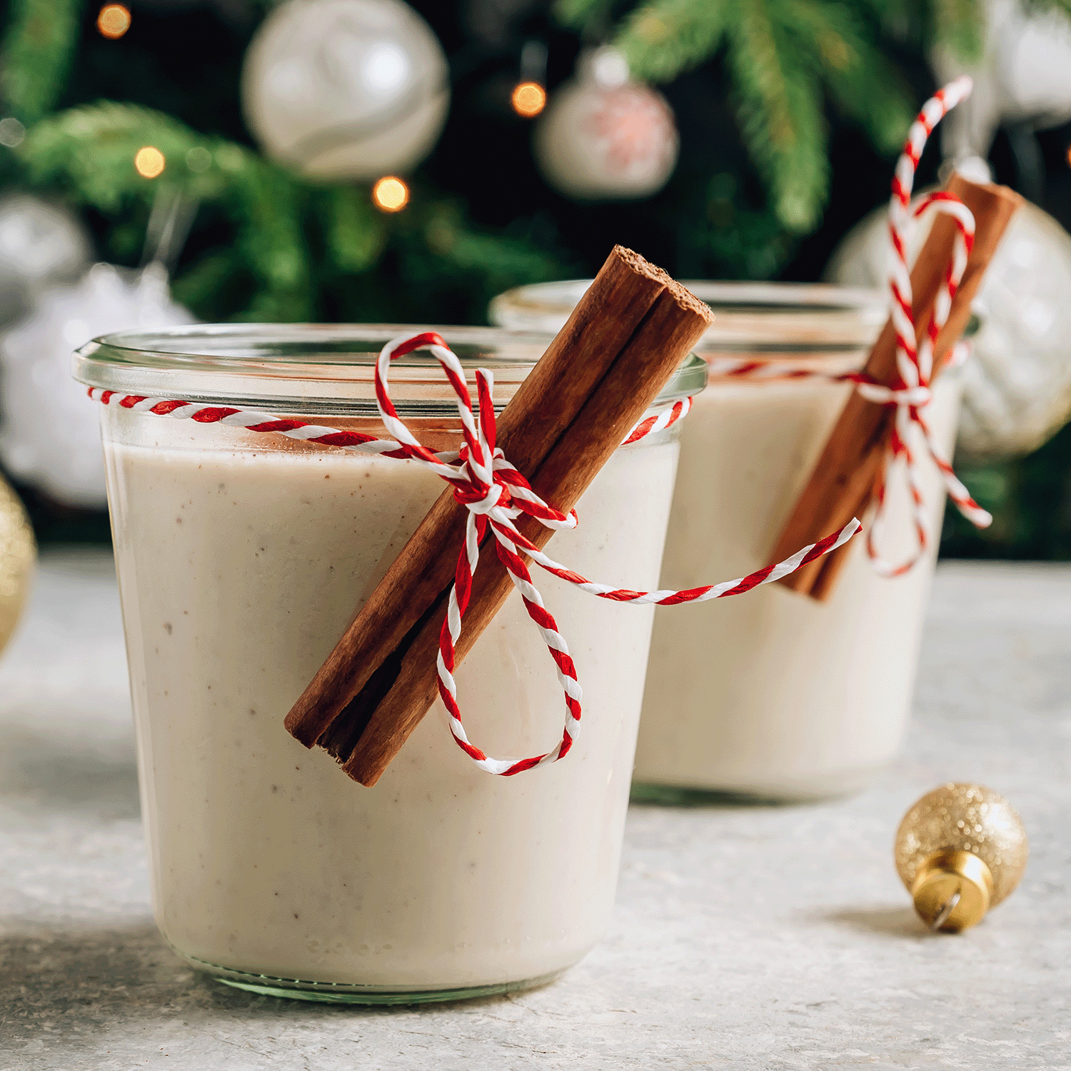 Two glass jars of eggnog garnished with cinnamon sticks and wrapped in red and white twine ribbons rest next to the Sleigh Ride Scented Jar Candle from the Tuscany Candle® SEASONAL Winter Flannels Collection. The candle casts a warm glow with its festive fragrance, while a beautifully adorned Christmas tree completes the holiday scene in the background.