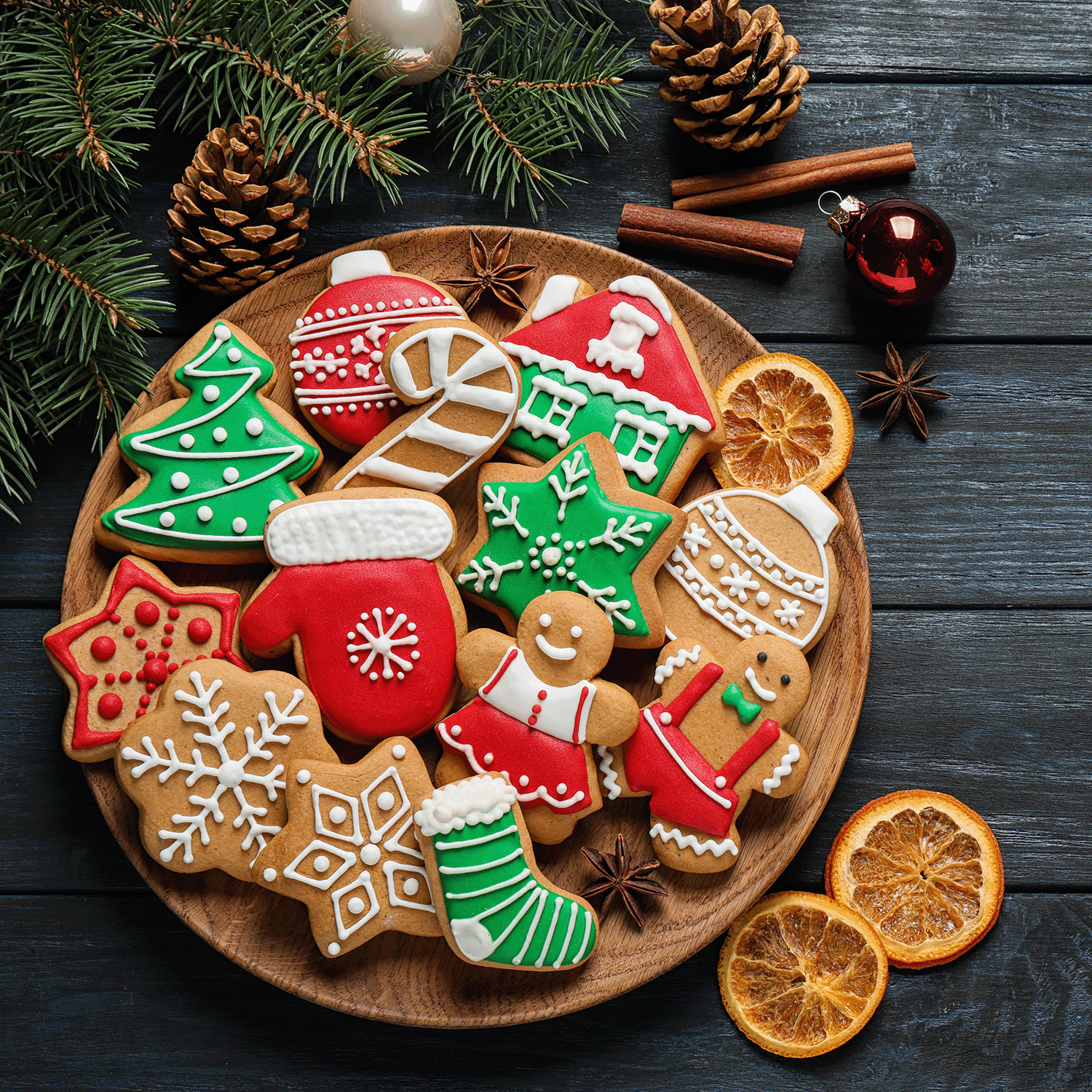 A wooden plate presenting an array of decorated Christmas cookies, such as gingerbread figures, mittens, and trees, is nestled amongst pine branches and pinecones. Nearby, the Christmas Cookies Scented Jar Candle (18 oz) from Tuscany Candle® SEASONAL's Winter Reverie Collection fills the air with its delightful baked vanilla fragrance.