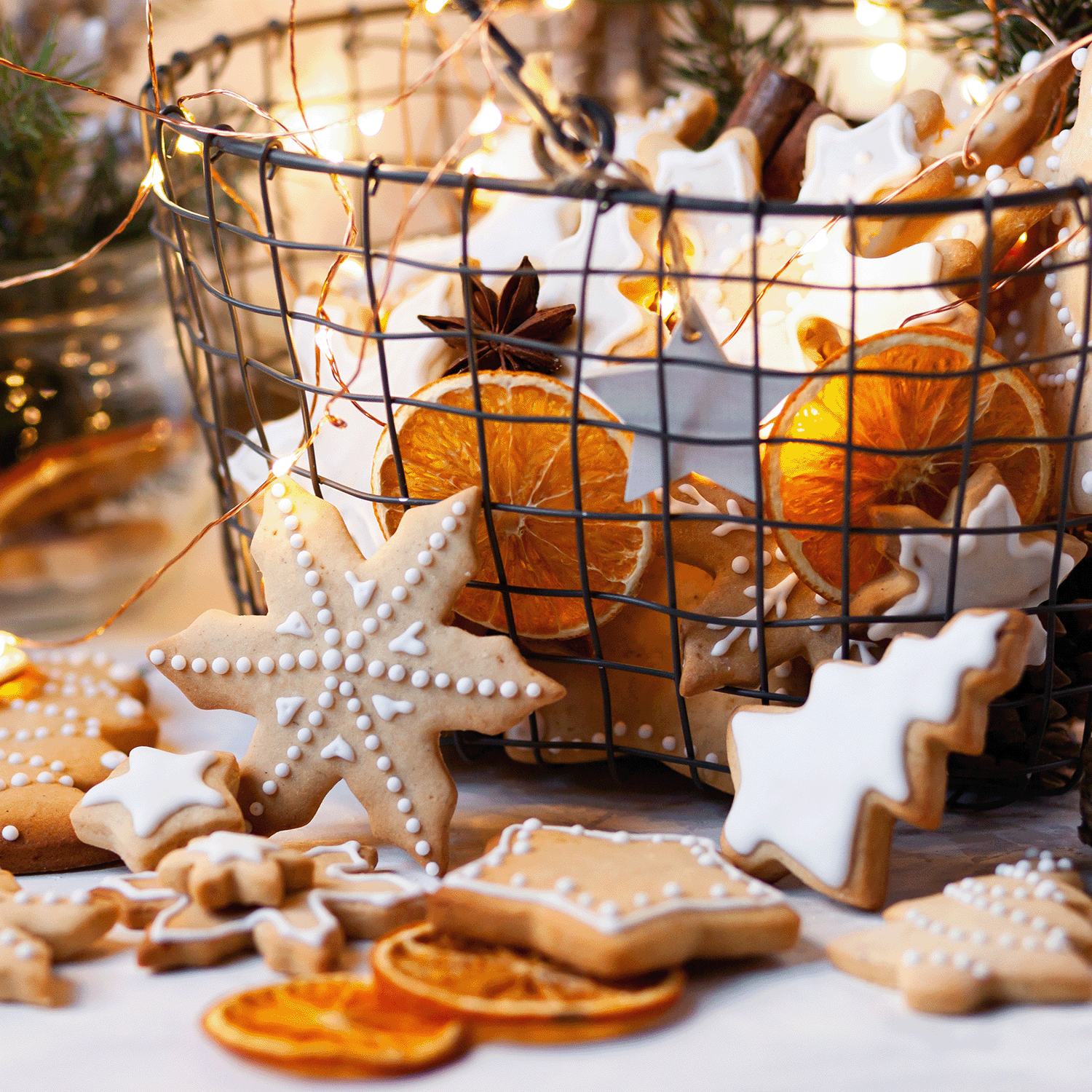 A wire basket filled with decorated gingerbread cookies and dried orange slices sits beside an Iced Gingerbread Scented Jar Candle (12 oz) by Tuscany Candle® SEASONAL. Cookies shaped like stars, trees, and snowflakes are arranged on a table with festive lighting, creating a cozy holiday atmosphere.