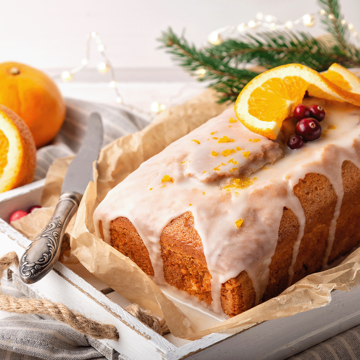 A glazed loaf cake adorned with orange slices and cranberries gleams on a tray, as the warm flicker of the Tuscany Candle® SEASONAL Iced Gingerbread Scented Jar Candle (12 oz) casts cozy shadows over the knife and string lights in the background.