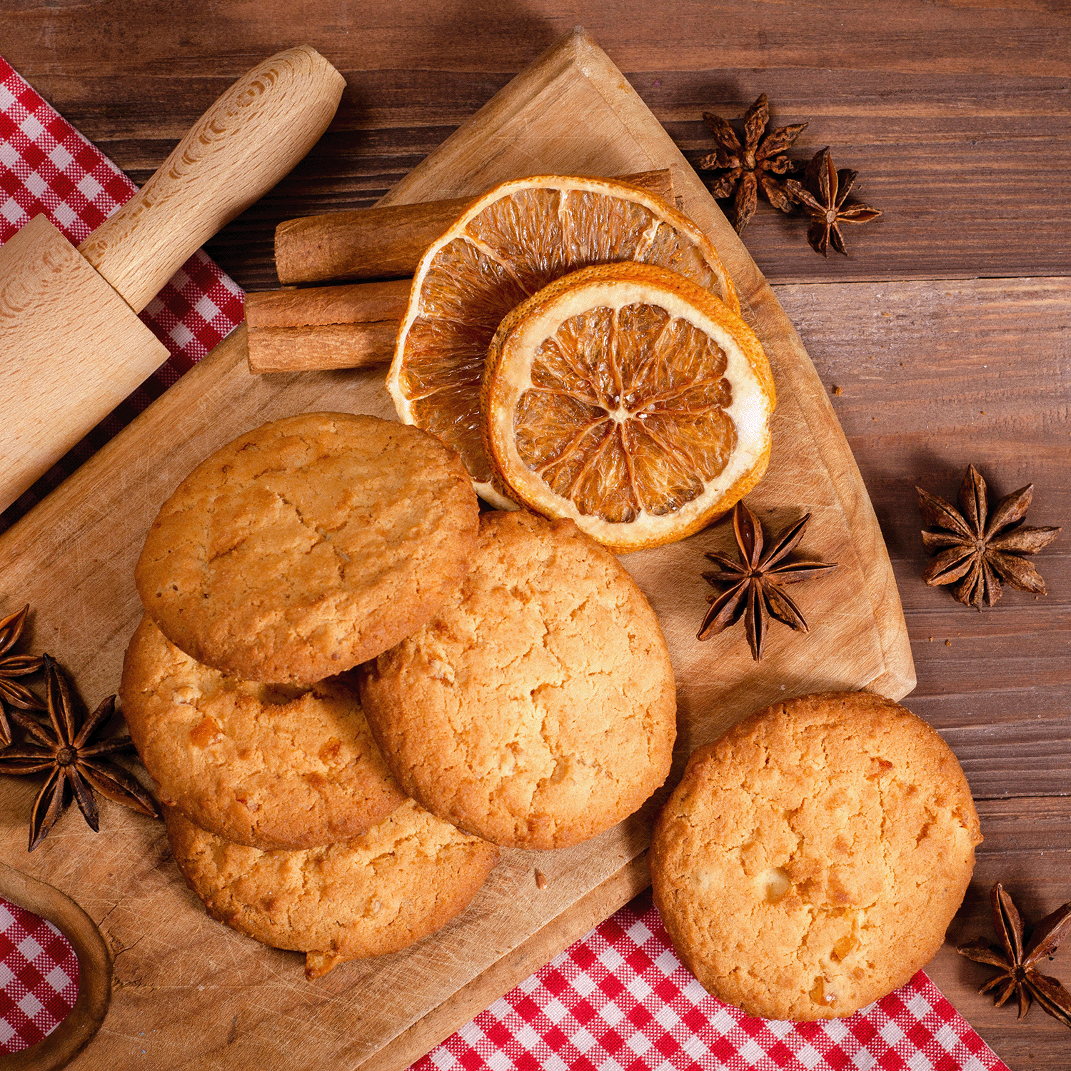 A wooden board with cookies, dried orange slices, spices, and a Tuscany Candle® SEASONAL Cranberry Shortbread: Sweet Cranberry & Sugar Scented Fall Candle (14 oz) rests on a wooden table with a red and white checkered cloth and a rolling pin.