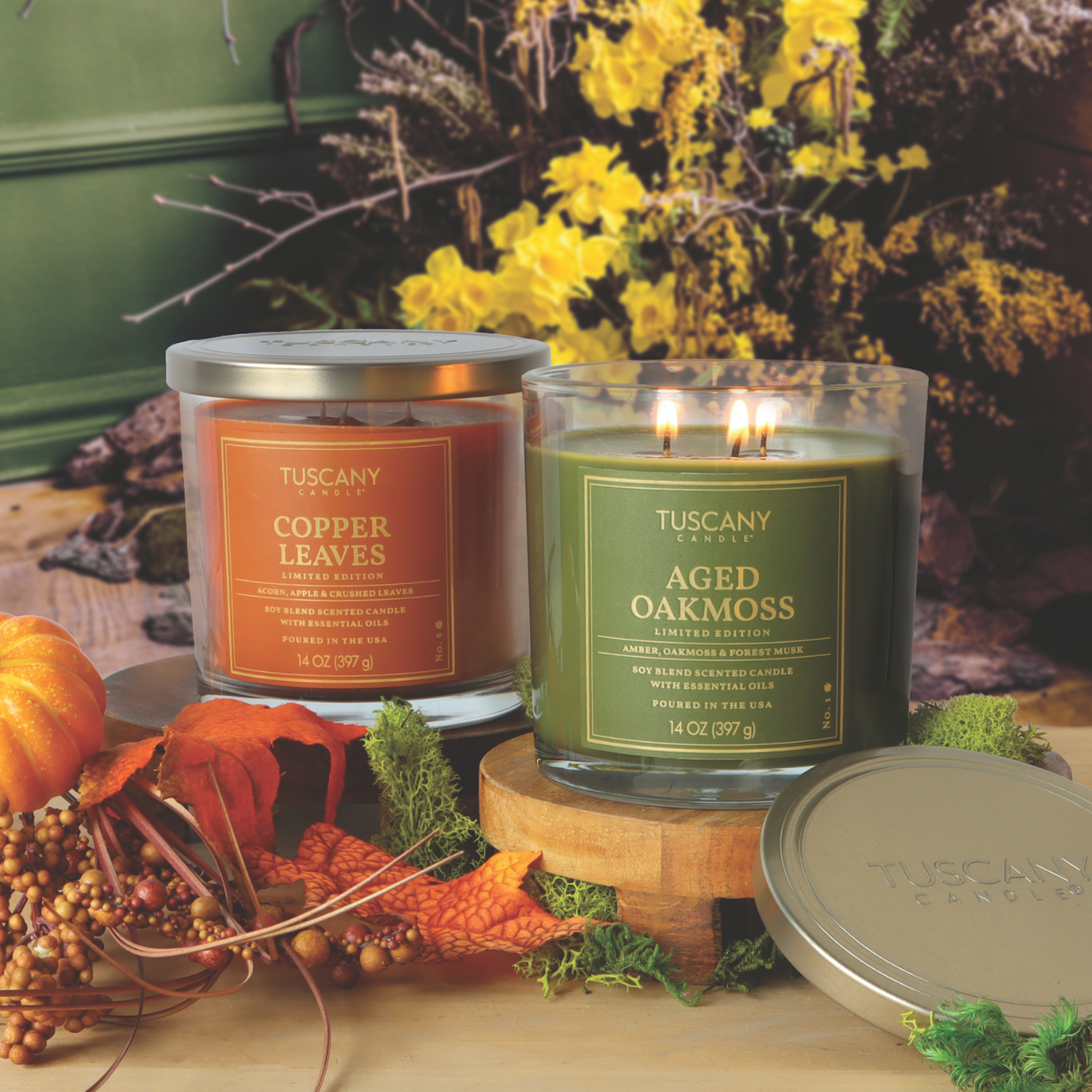 Two Tuscany Candle® SEASONAL 14 oz candles—Copper Leaves and Aged Oakmoss: Forest Musk & Amber—are set on a table with autumn-themed décor and a floral arrangement in the background.