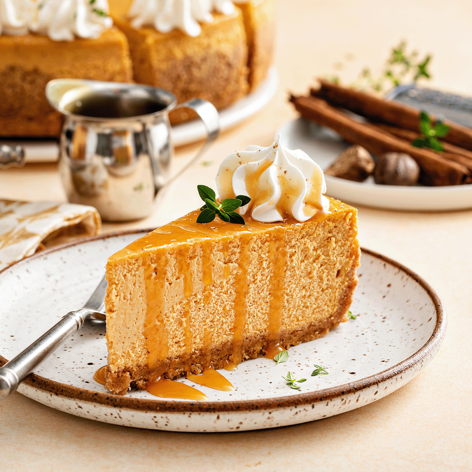 A slice of vanilla cheesecake with whipped cream and caramel drizzle sits on a plate, surrounded by a fork, sauce pitcher, spices, and the warm glow of Tuscany Candle® SEASONAL’s Simply Fall: Spiced Pumpkin & Cheesecake Scented Candle (14 oz).
