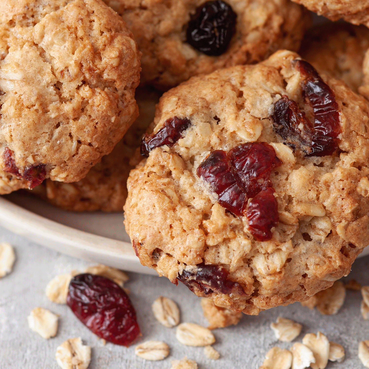 Oatmeal cookies with dried cranberries on a plate, surrounded by oats—the perfect treat to enjoy with the warm glow of Tuscany Candle® SEASONAL Autumn Oatmeal Cookie: Harvest Spices Scented Fall Candle from the Bakery Tins Collection (12 oz).